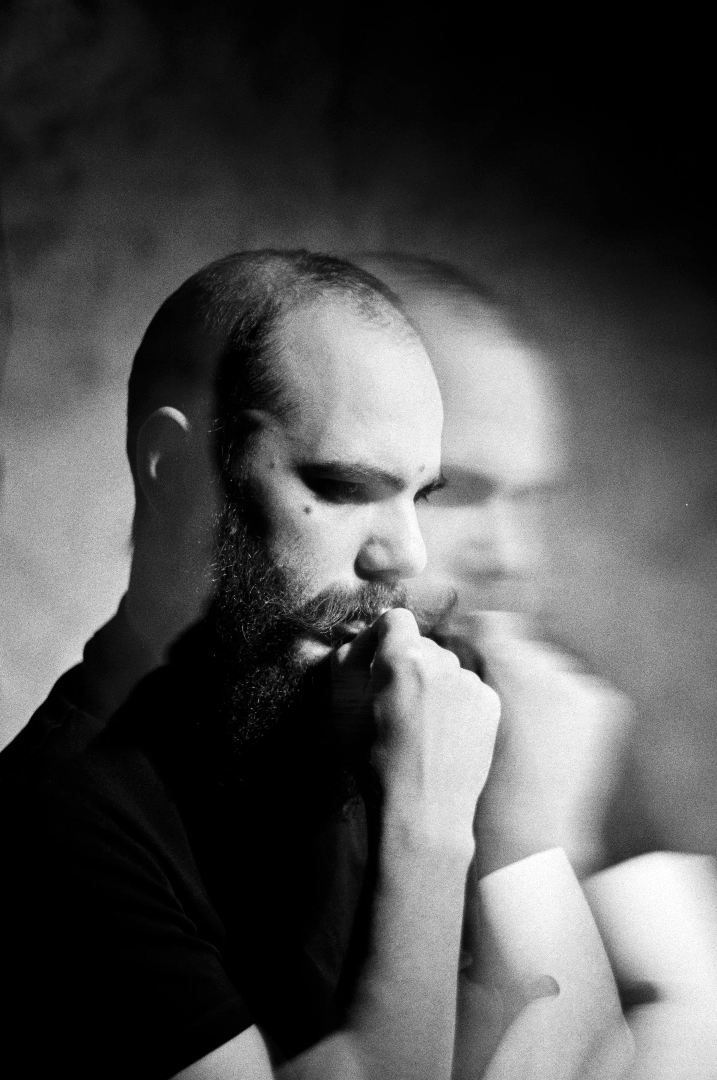 Black and white portrait of a man with a beard, looking down thoughtfully with his hand near his mouth, with a blurred duplicate of his face in the background.