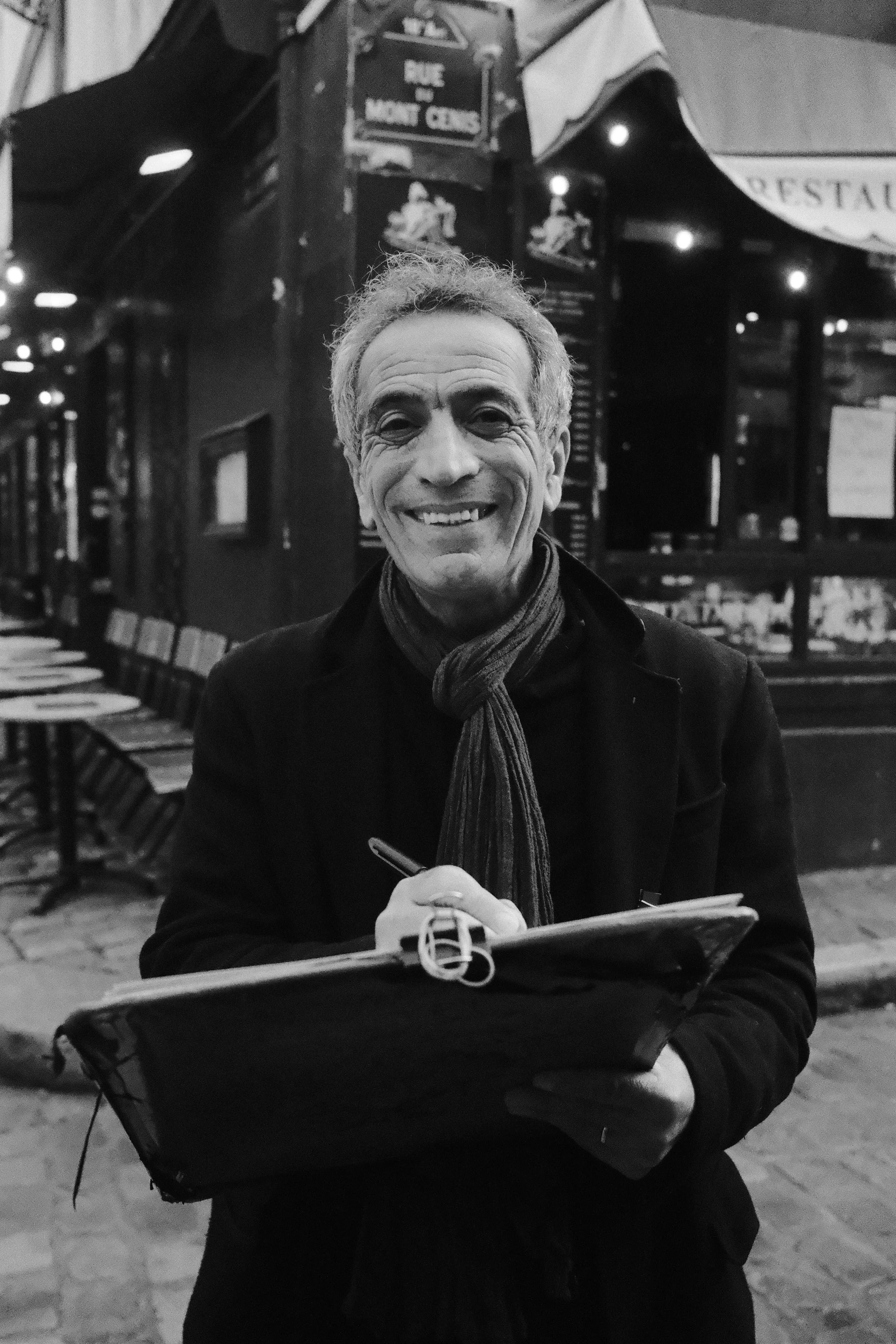 A smiling man with gray hair holding a notebook and pen on a street at night in front of a restaurant with chairs and tables outside.