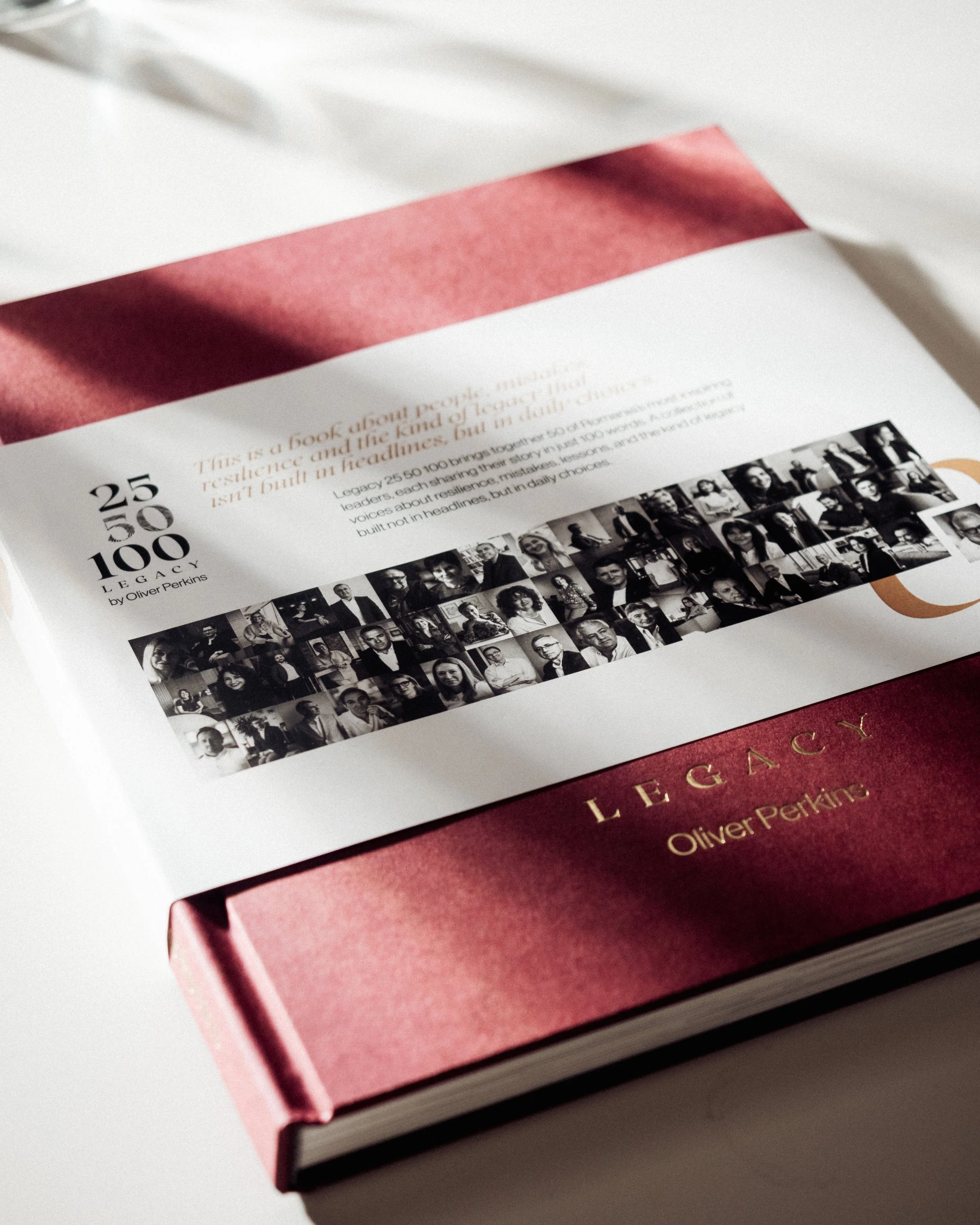 A hardcover book titled "Legacy" by Oliver Perkins with a red cover and gold lettering, with a paper band on top featuring black and white portraits of various people.