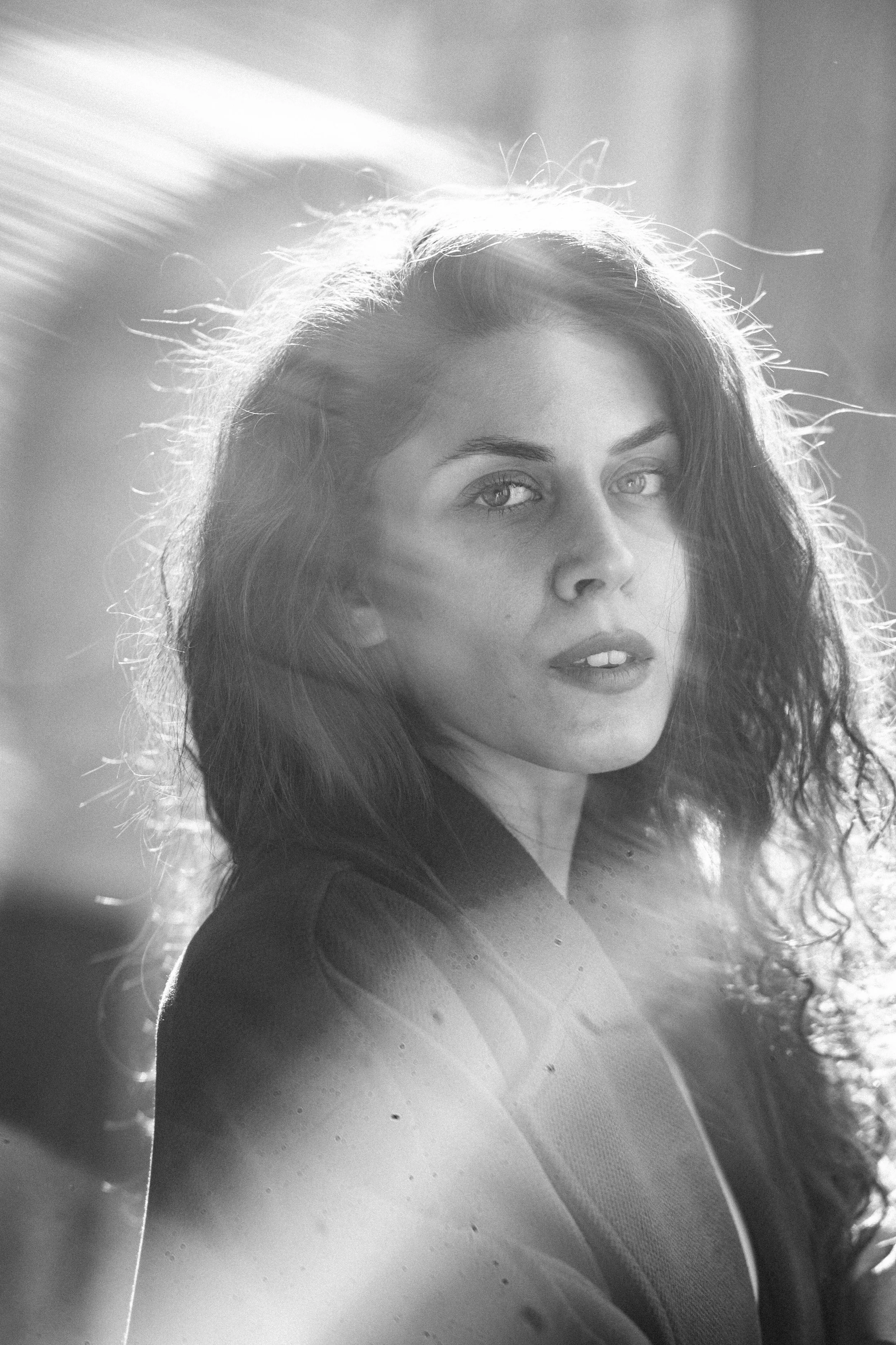 A black and white photo of a woman with curly hair, looking towards the camera with sunlight highlighting her hair and face.
