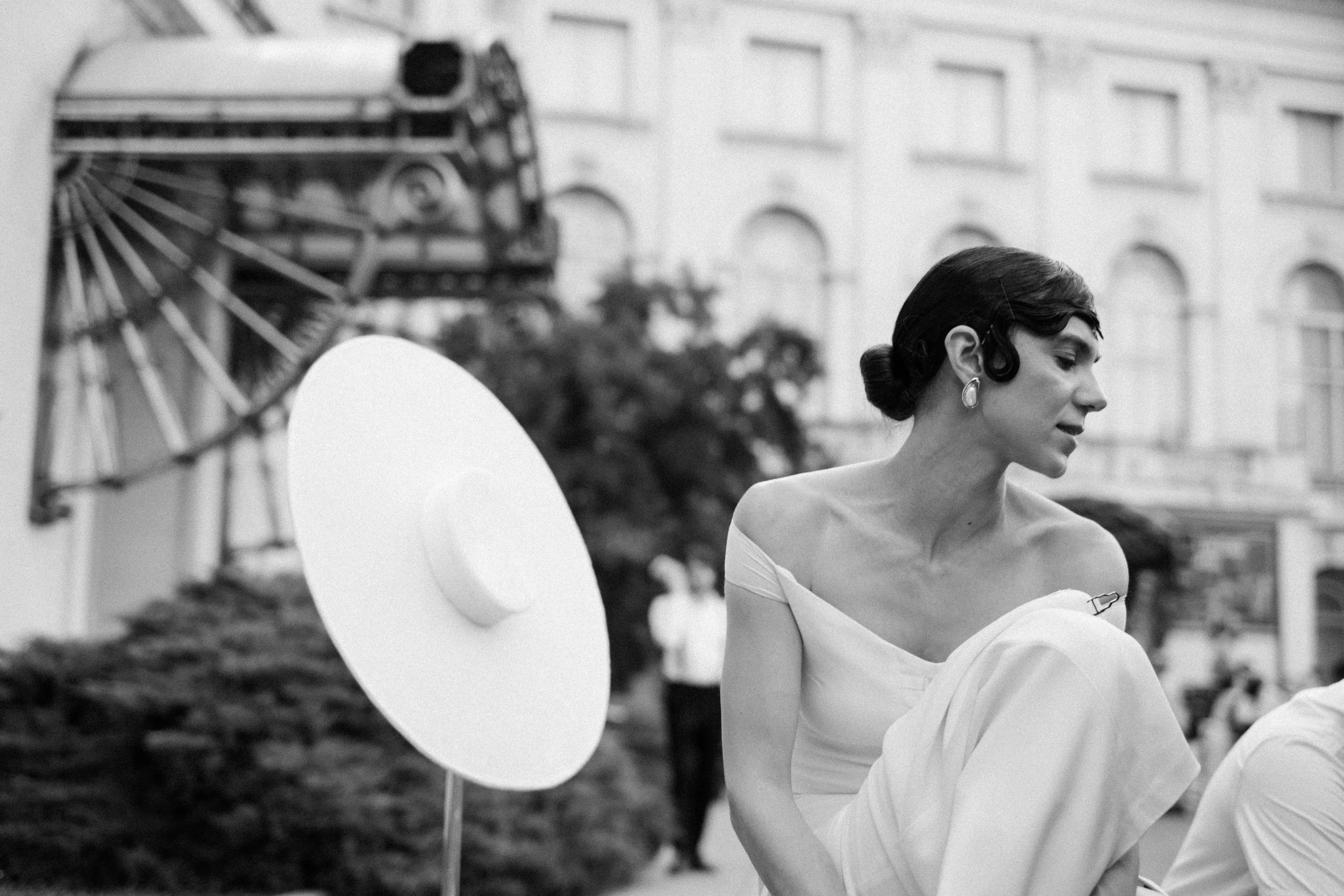 A black and white photo of a woman wearing an off-shoulder dress and large earrings, standing outdoors next to a large parasol, with a Ferris wheel and a building with arched windows in the background.