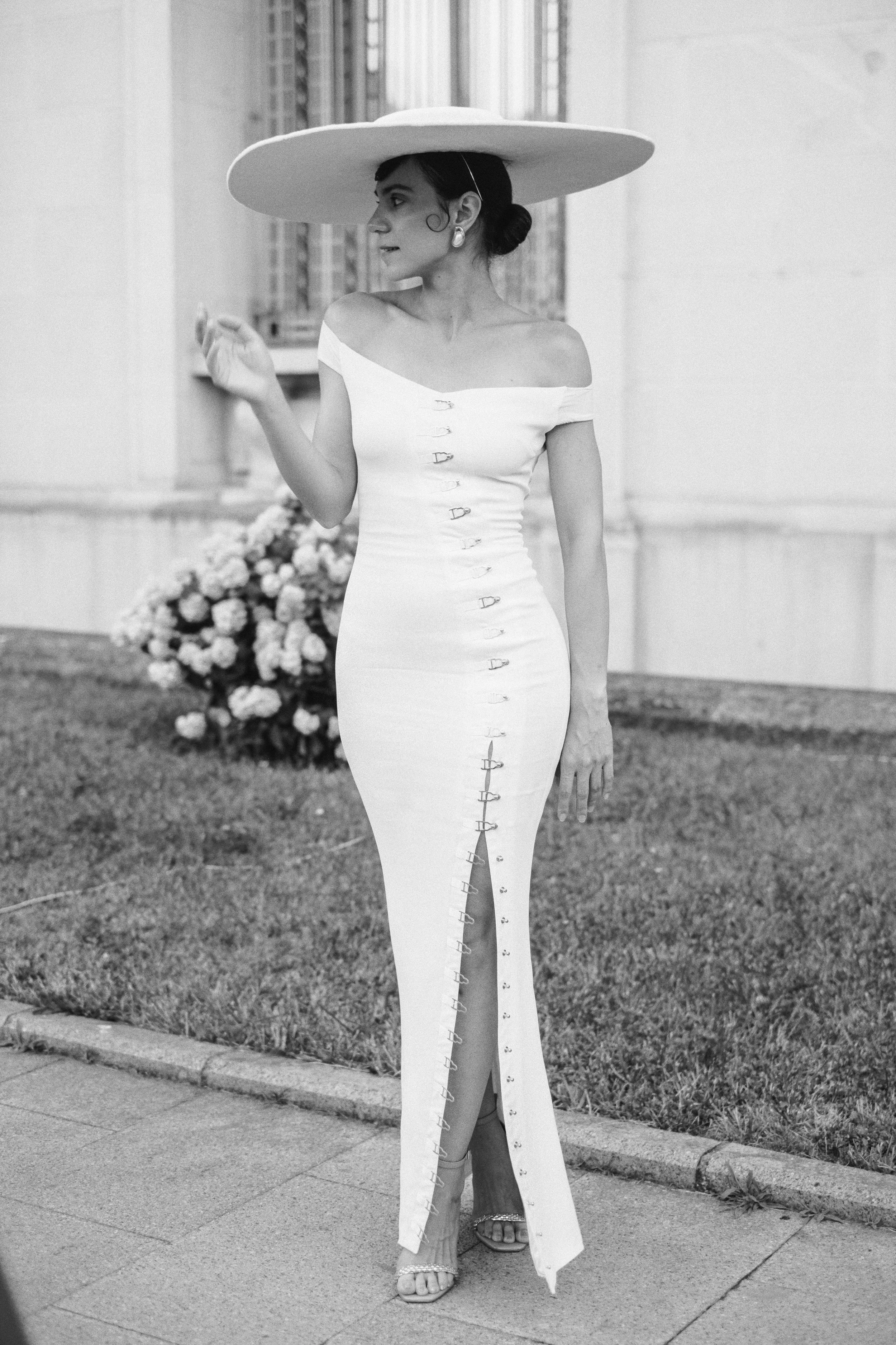 A woman dressed in a long, elegant white dress with a slit, wearing a large wide-brimmed hat, standing outdoors near a building with flowers in the background.