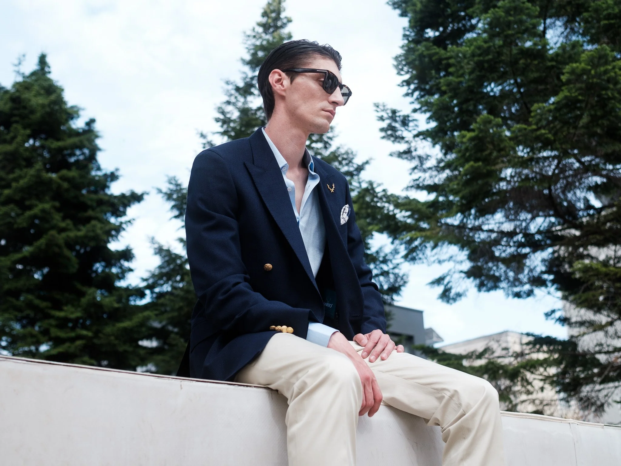 Man in sunglasses and blazer sitting outdoors near trees.