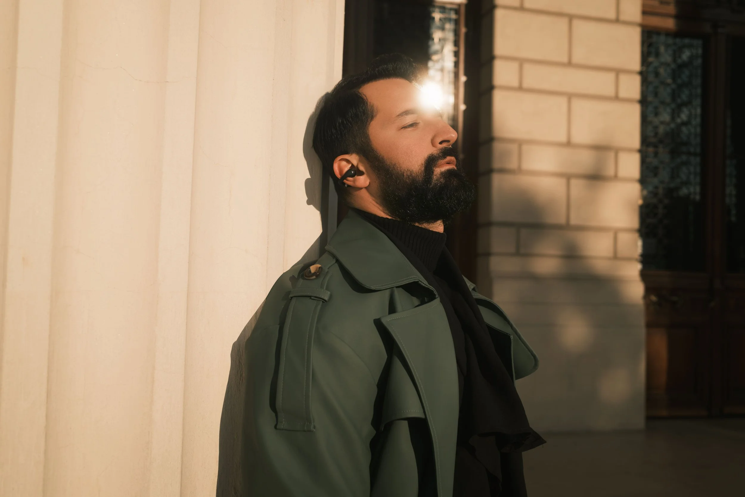 Ahmed Hatem leans against a stone wall with his eyes closed, wearing a green trench coat and earphones, sunlight falling across his face.