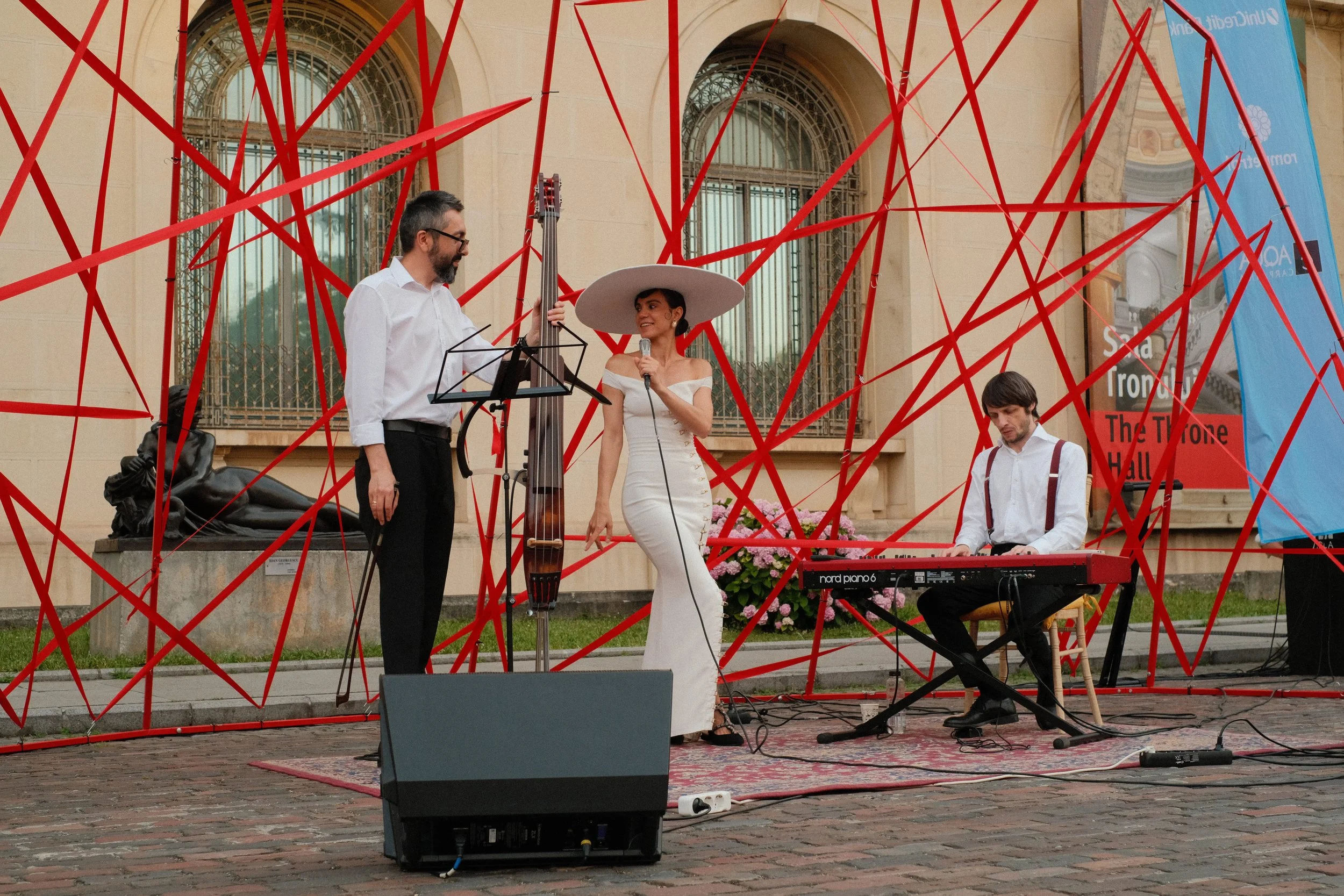 Musicians performing outdoors in front of a historic building with arched windows. A man is playing a double bass, a woman is singing with a microphone, and another man is playing a keyboard. Red string art installation surrounds the performers.
