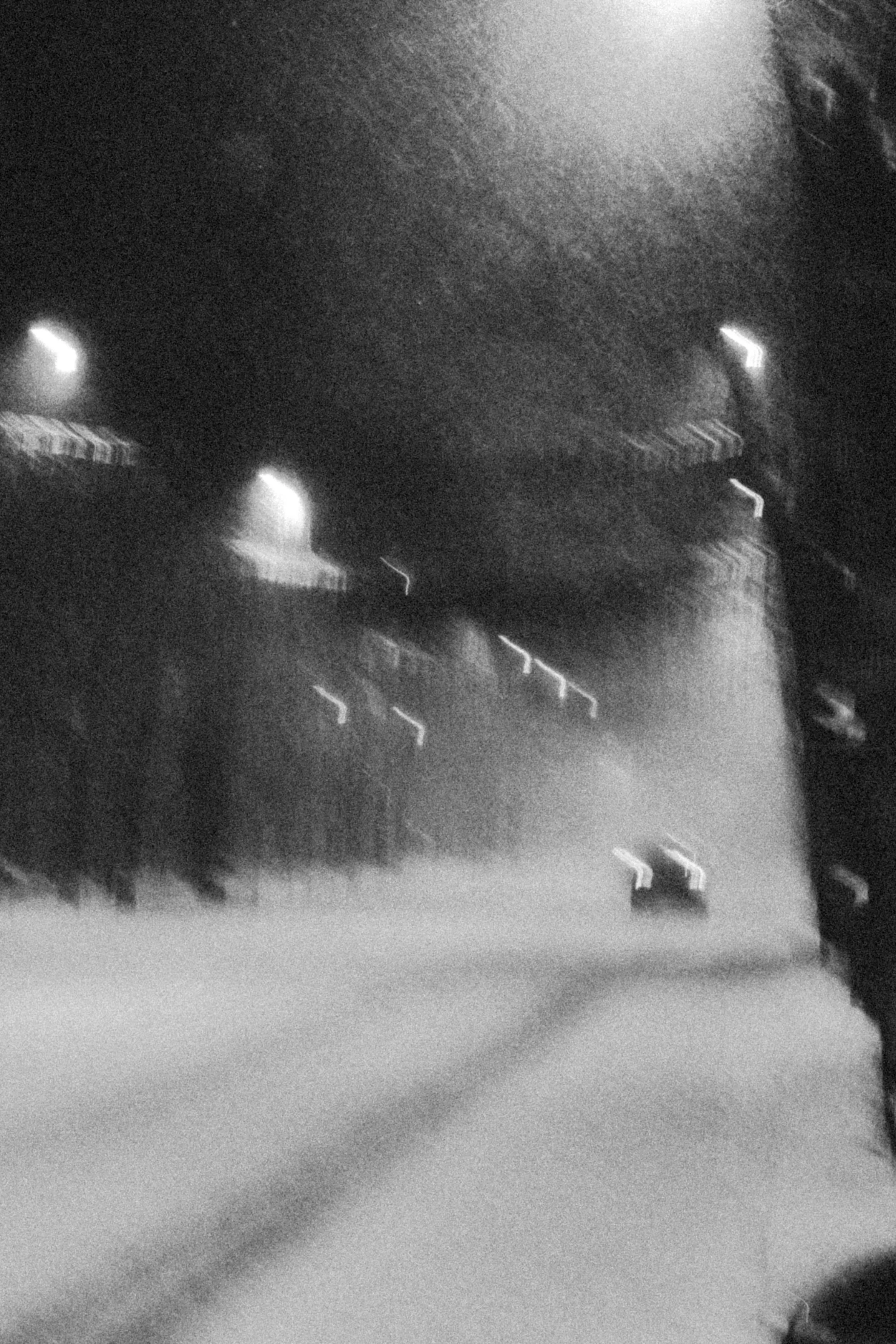 A blurry black-and-white photo of a dark street at night with streetlights and some buildings in the background.