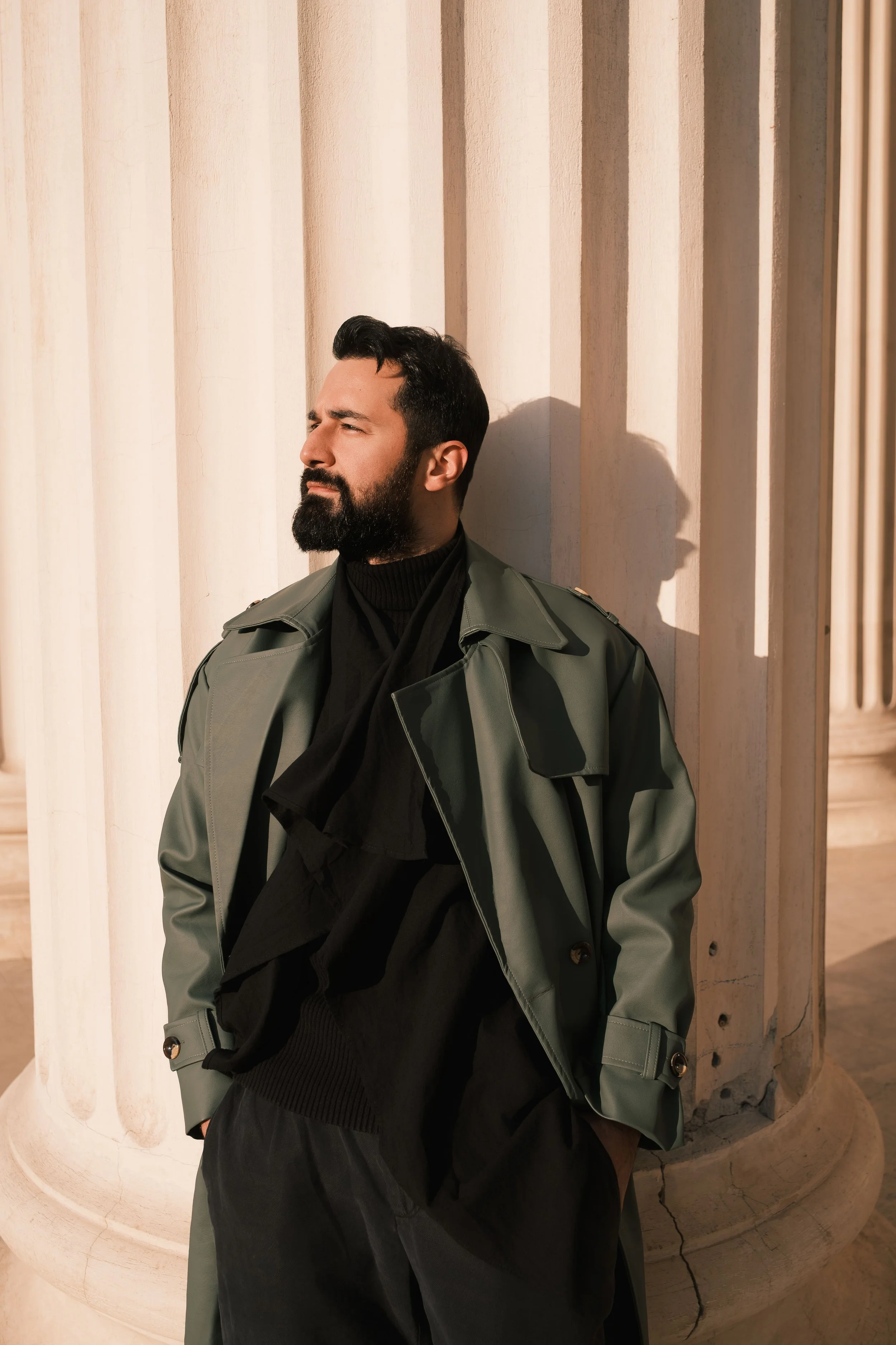Ahmed Hatem stands against a fluted column of a neoclassical building, wearing a gray trench coat and black turtleneck, looking to the side as his shadow stretches across the stone.