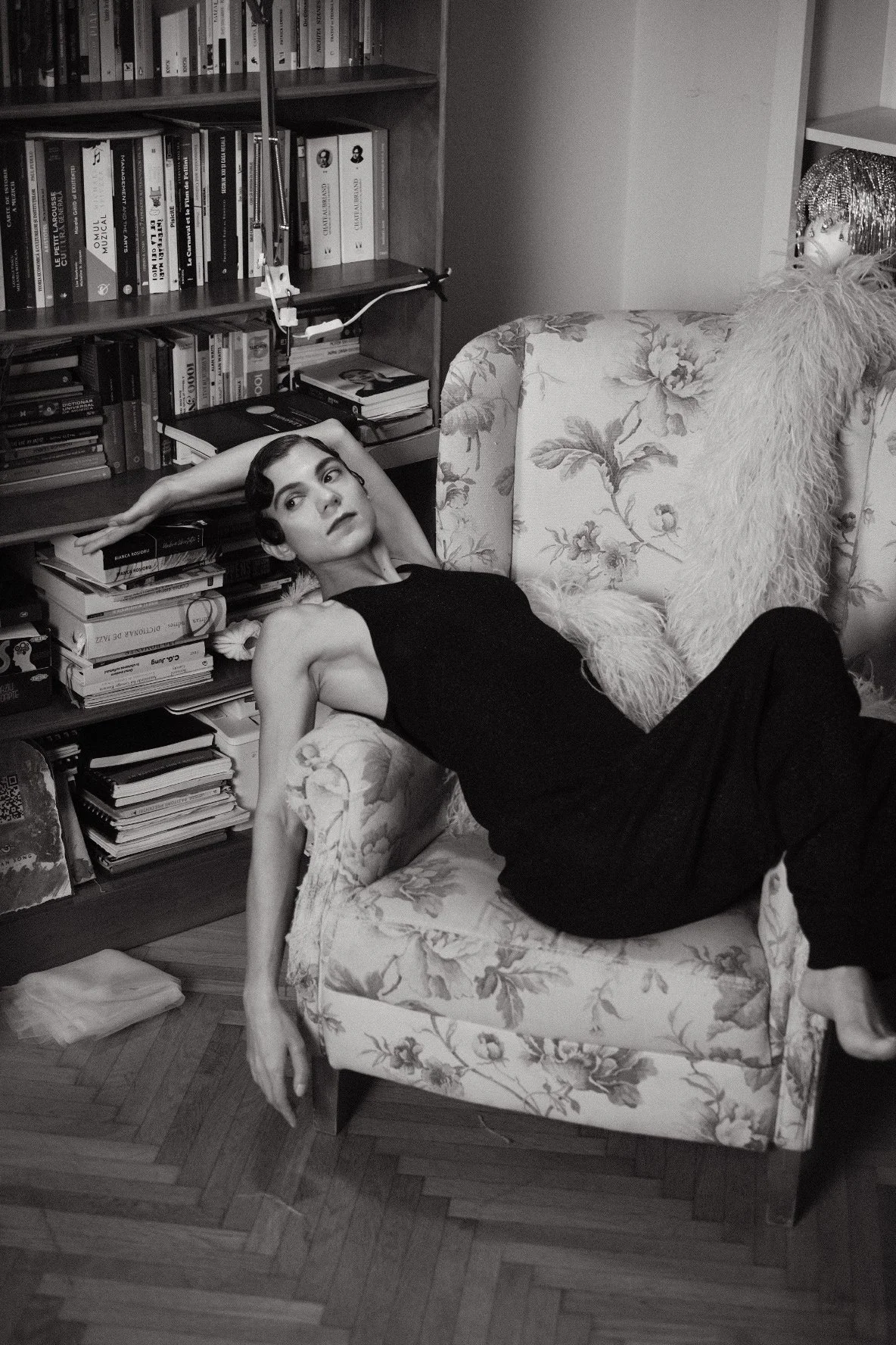 Black and white portrait of Sorina Rotaru reclining in a floral armchair next to a bookshelf, Bucharest home