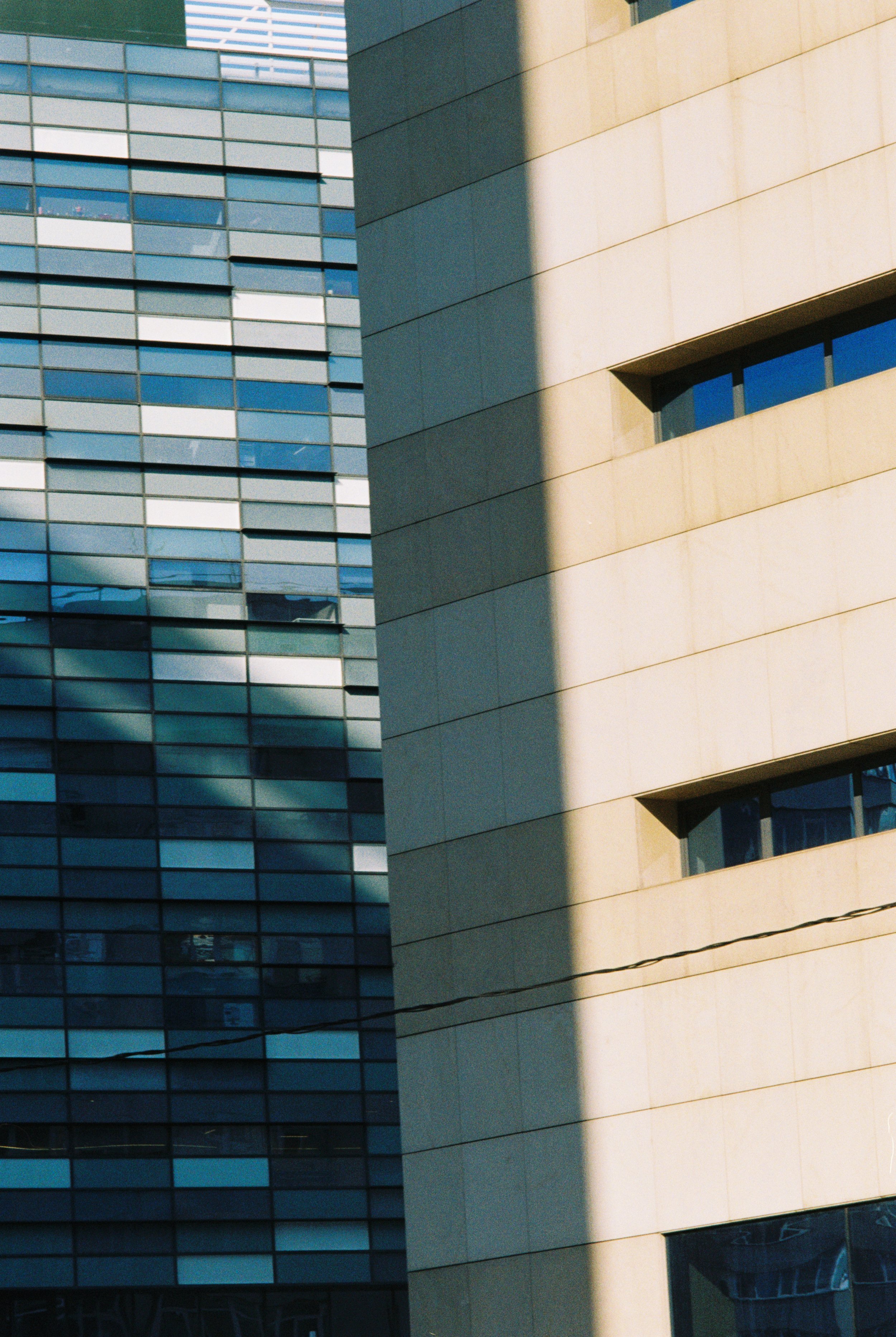 Two modern high-rise buildings with glass and beige exterior walls, partially shaded by nearby structures, under a clear sky.