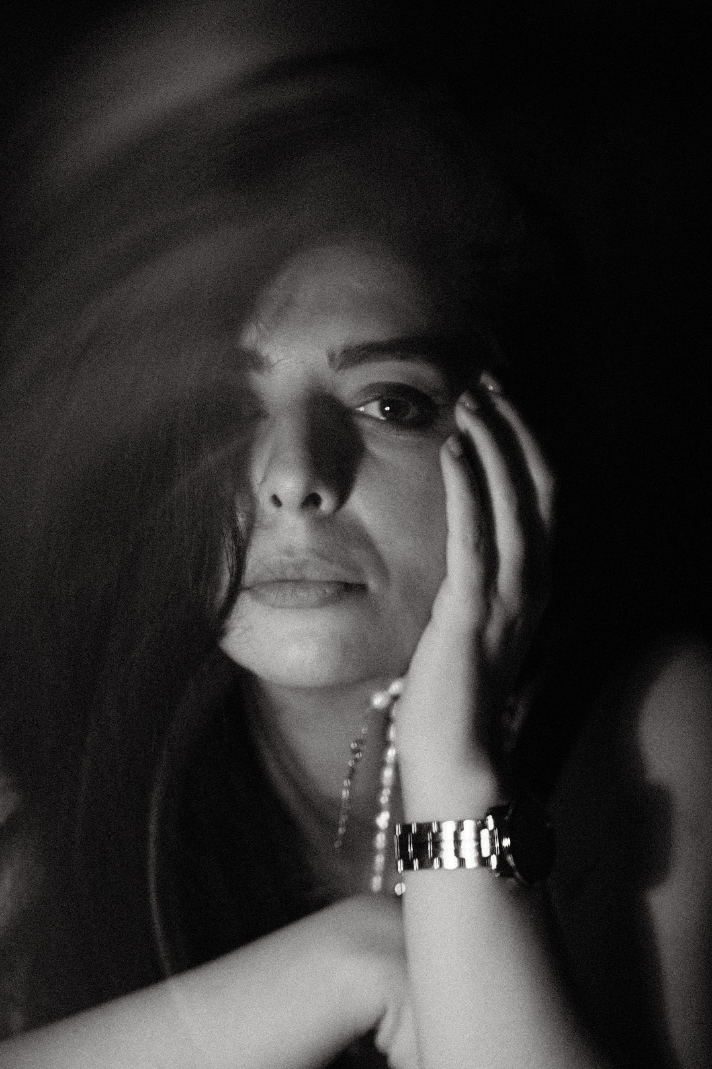 A black and white close-up photo of a woman with long hair, wearing jewelry including a watch and a bracelet, with her hand near her face, partially covering her eye.