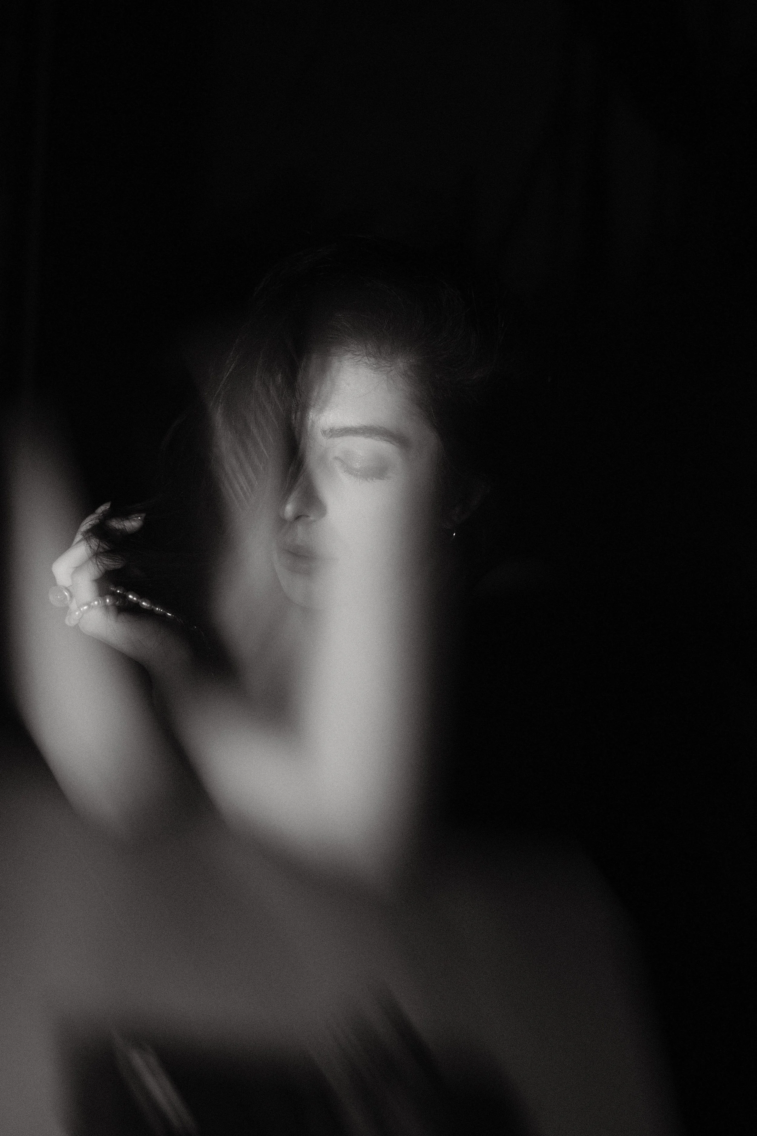 Black and white portrait of a woman with closed eyes, wearing earrings and a pearl necklace, with her hand near her head, against a dark background.
