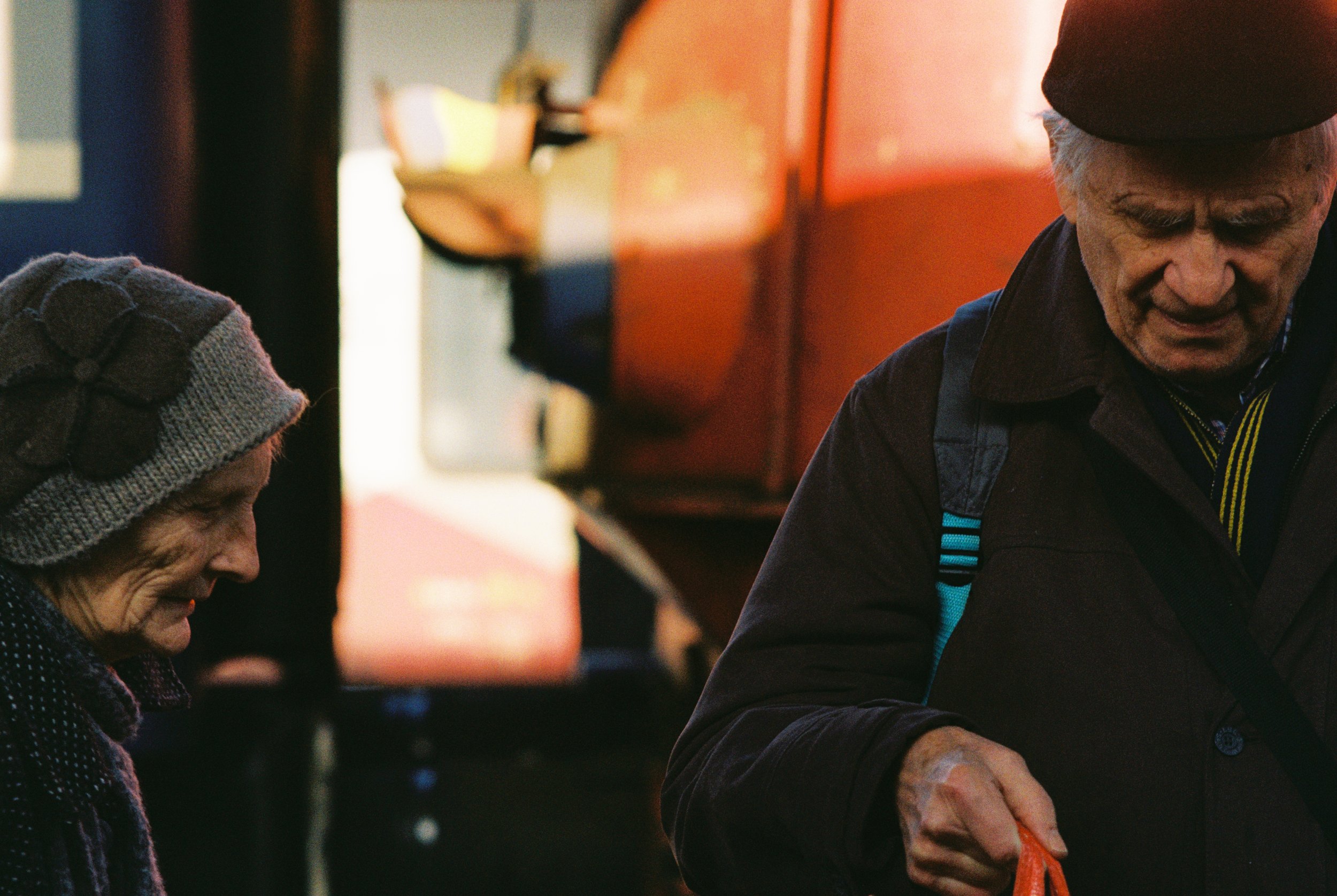 An elderly woman wearing a knit hat and a dark coat, smiling and looking down, standing next to an elderly man in a dark jacket and a beret, who is looking down and holding an orange object, possibly a shopping bag, with a blurry urban background.
