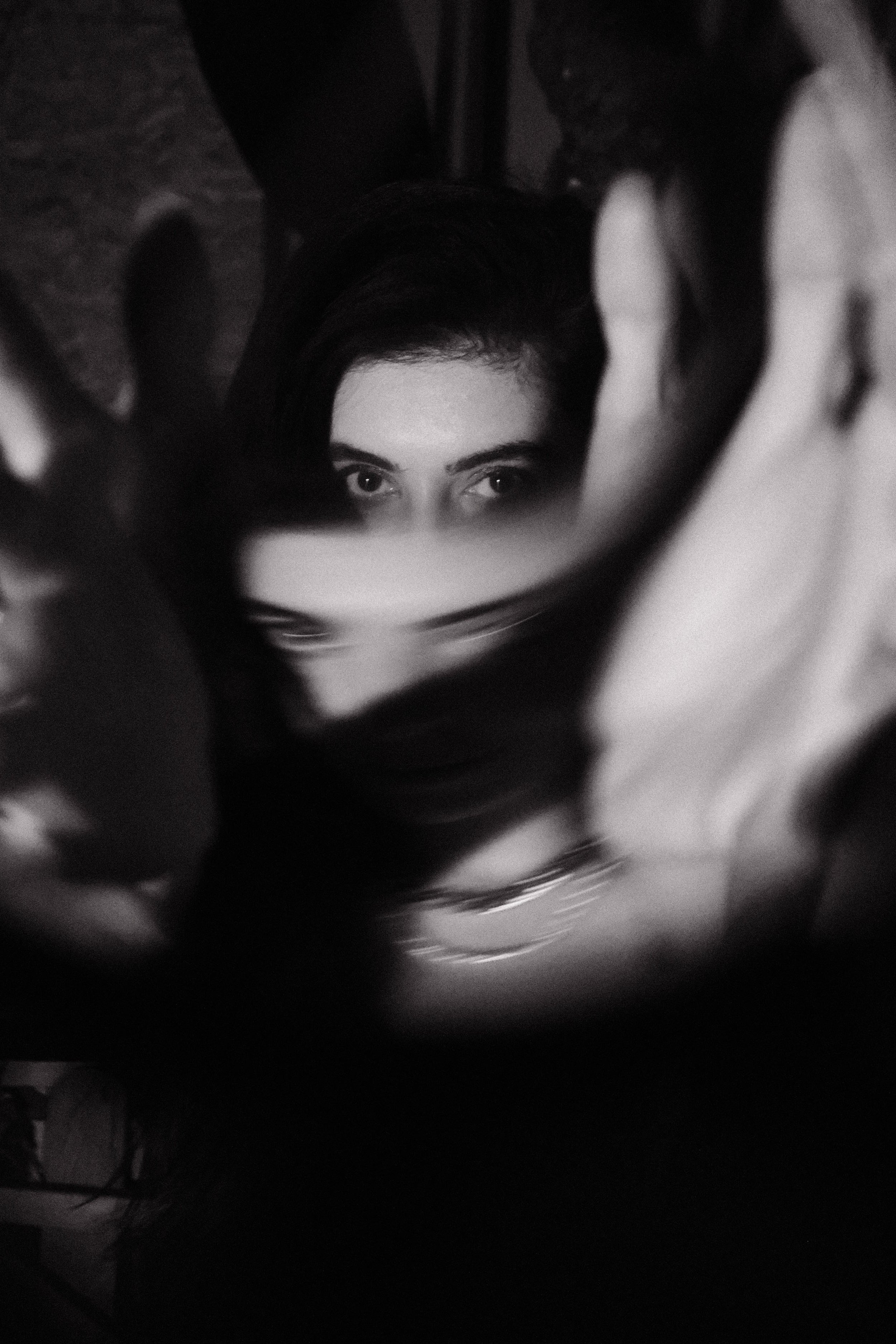 A black and white photo of a woman with dark hair and eyes, partially obscured by a reflective surface or object in front of her.