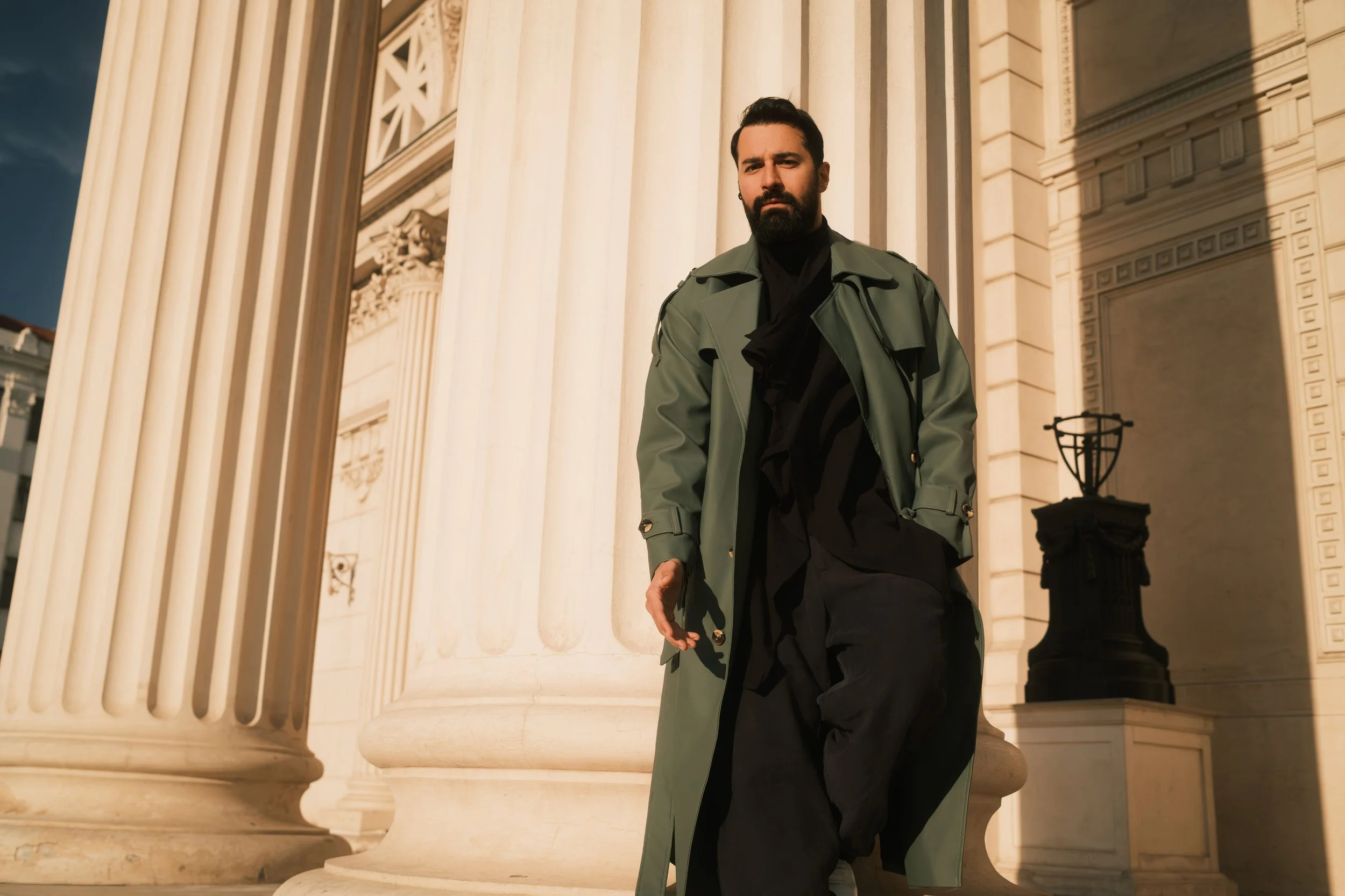 Ahmed Hatem walks past a neoclassical facade in Bucharest, wearing a green trench coat, captured in a quiet, in-between moment.