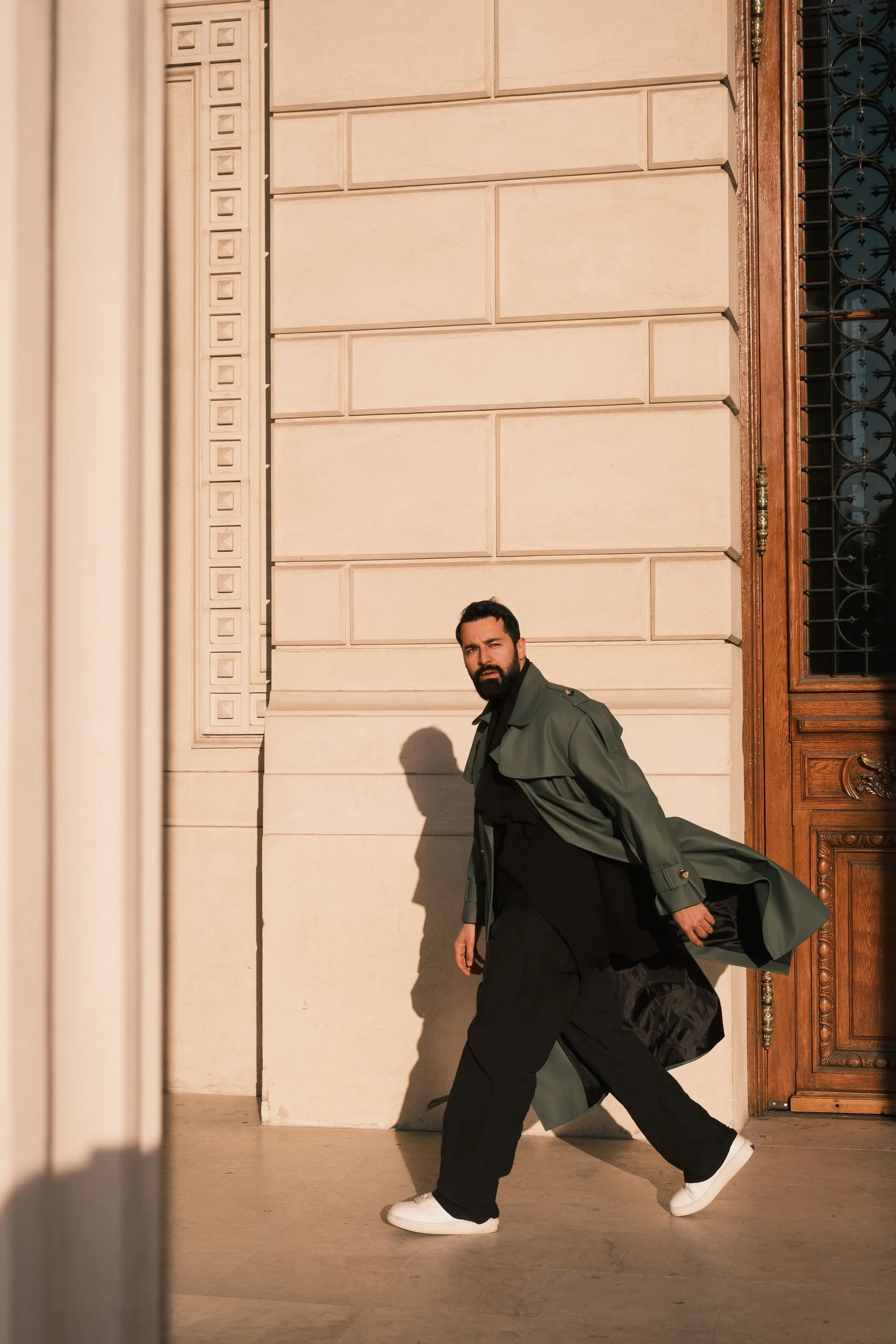 Ahmed Hatem walks alongside a textured stone wall and an ornate wooden door, wearing a green trench coat and white sneakers, blending classical architecture with a modern presence.