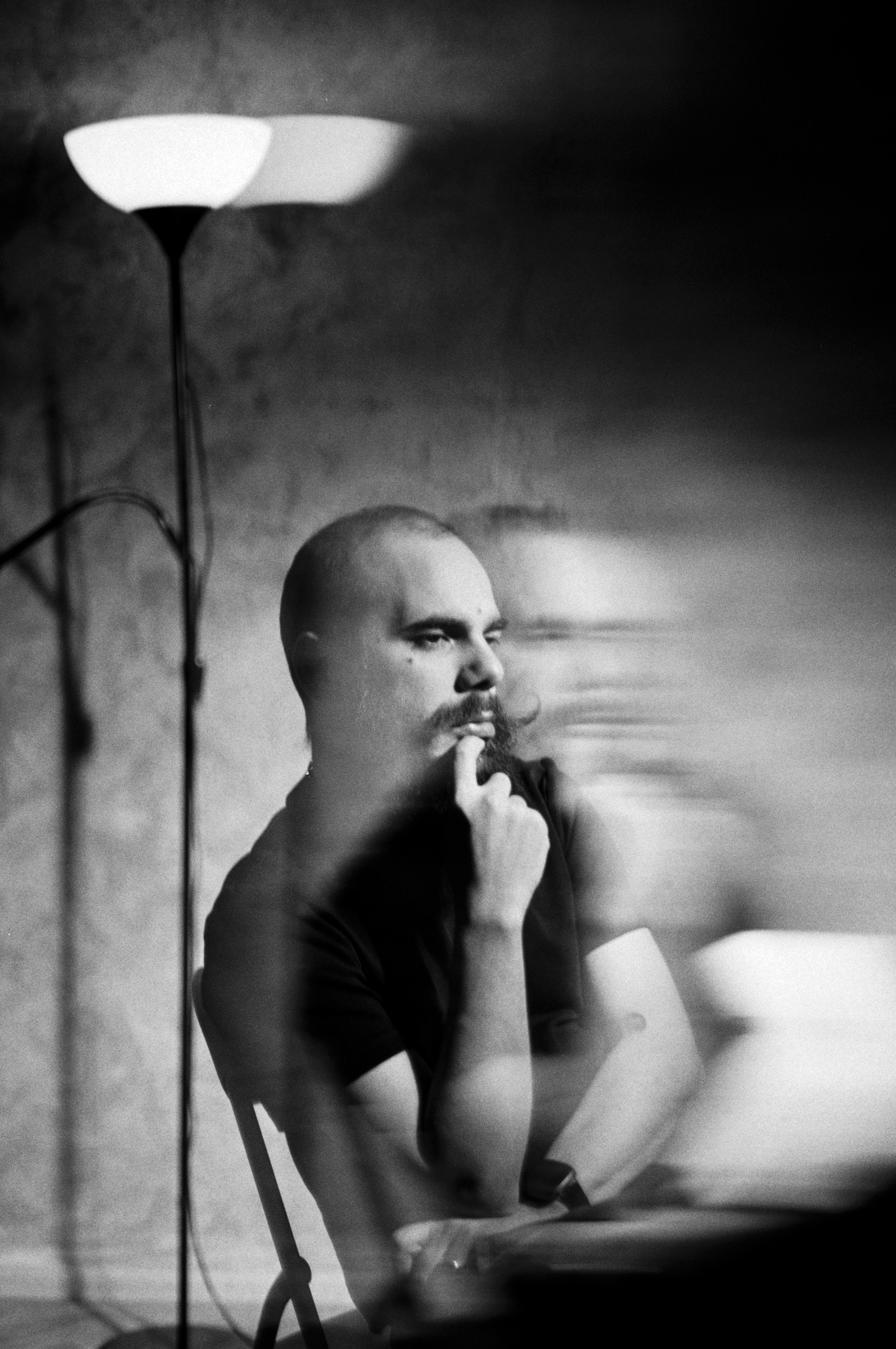 A black-and-white photograph of a man with a shaved head and beard, sitting at a table, with a contemplative expression, resting his chin on his finger, and a blurry reflection or motion effect to the right, next to a tall lamp.