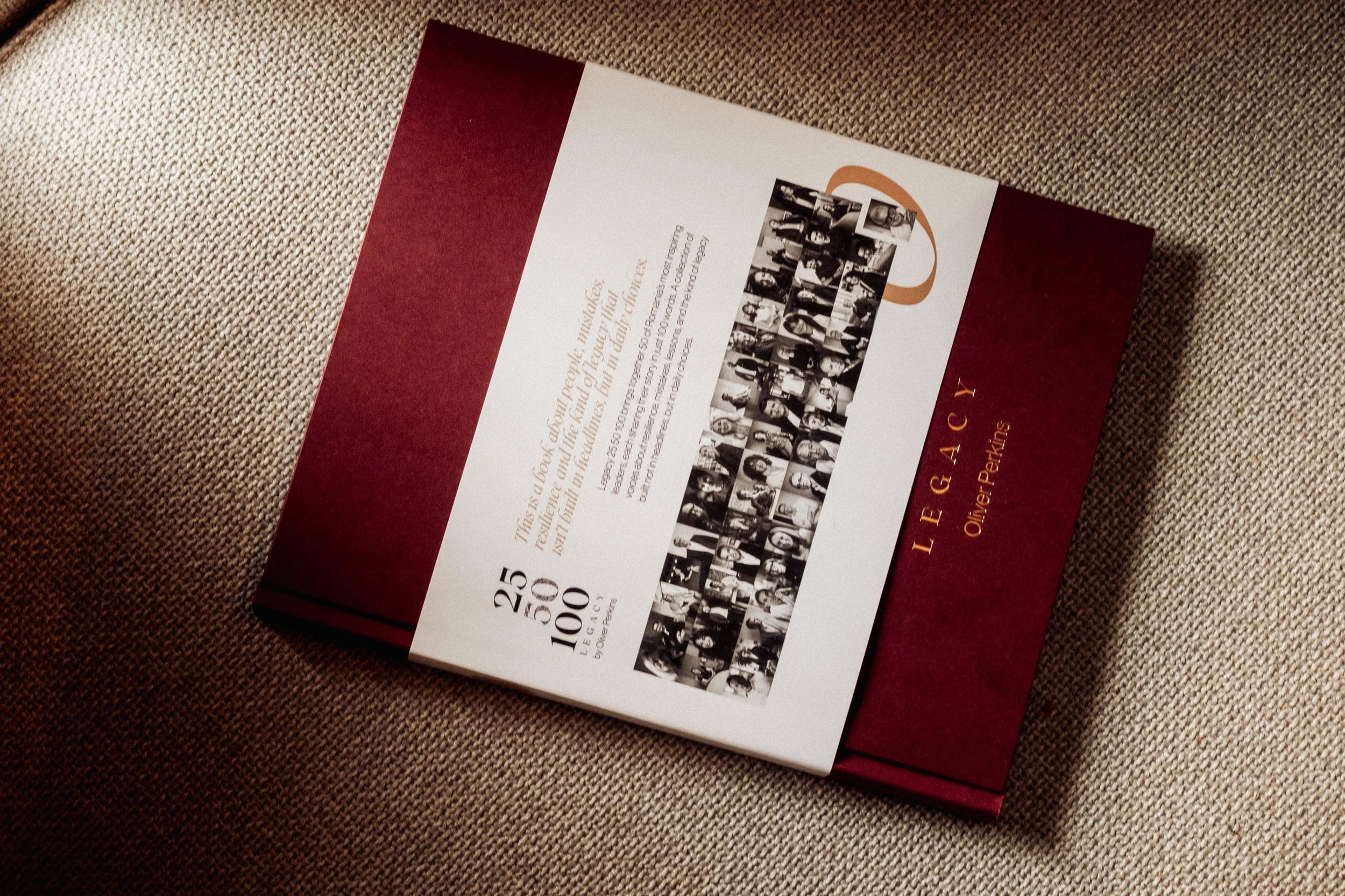 A hardcover book titled "Legacy" by Oliver Perks lying on a beige fabric surface. The book has a burgundy cover with a white central label displaying a collage of black and white photographs of people, and some text about the book inside.
