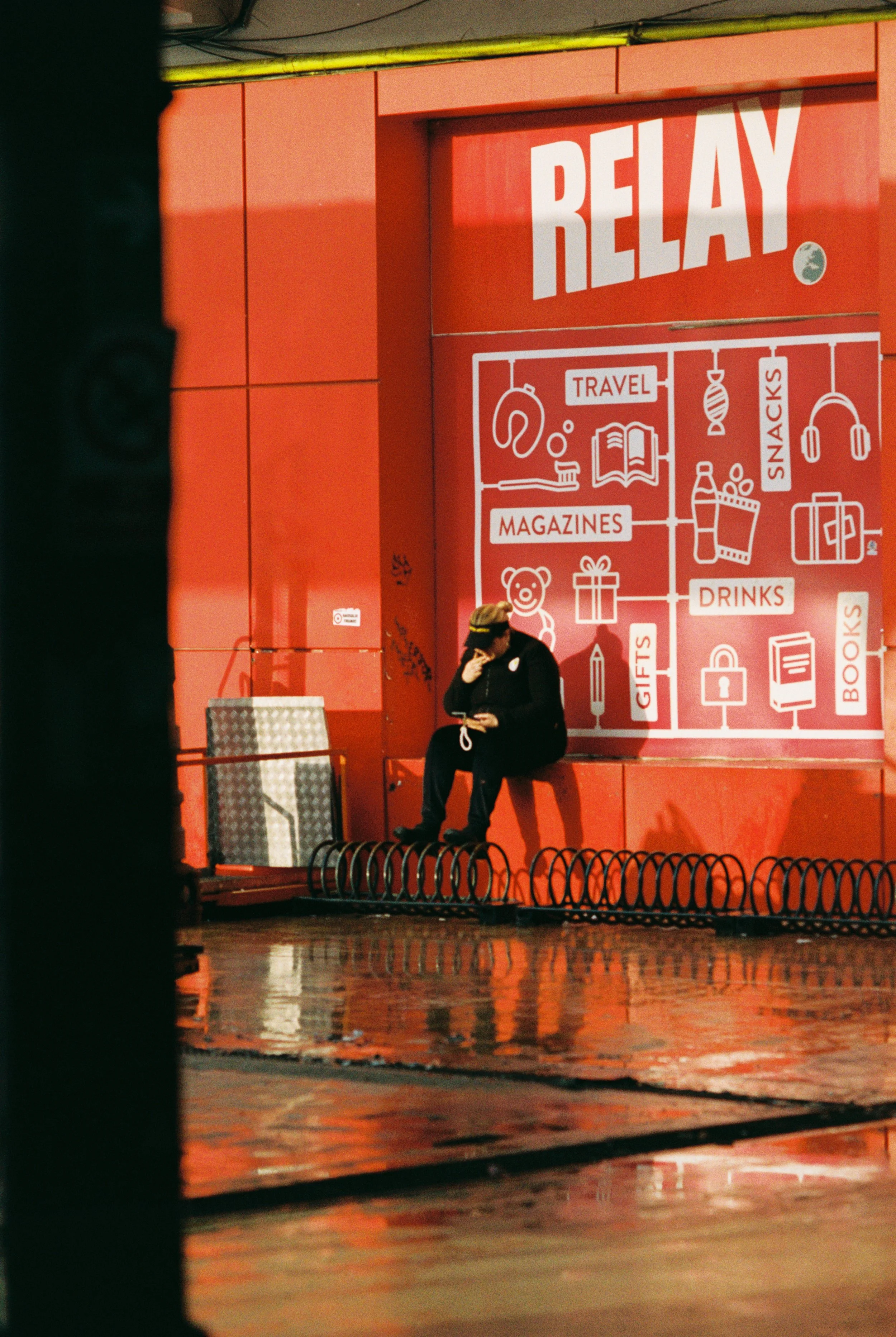 A person sitting on a bench in front of a red wall with white and orange signage advertising a convenience store. The signage indicates sections for travel, magazines, gifts, drinks, snacks, and books, with icons representing each category.