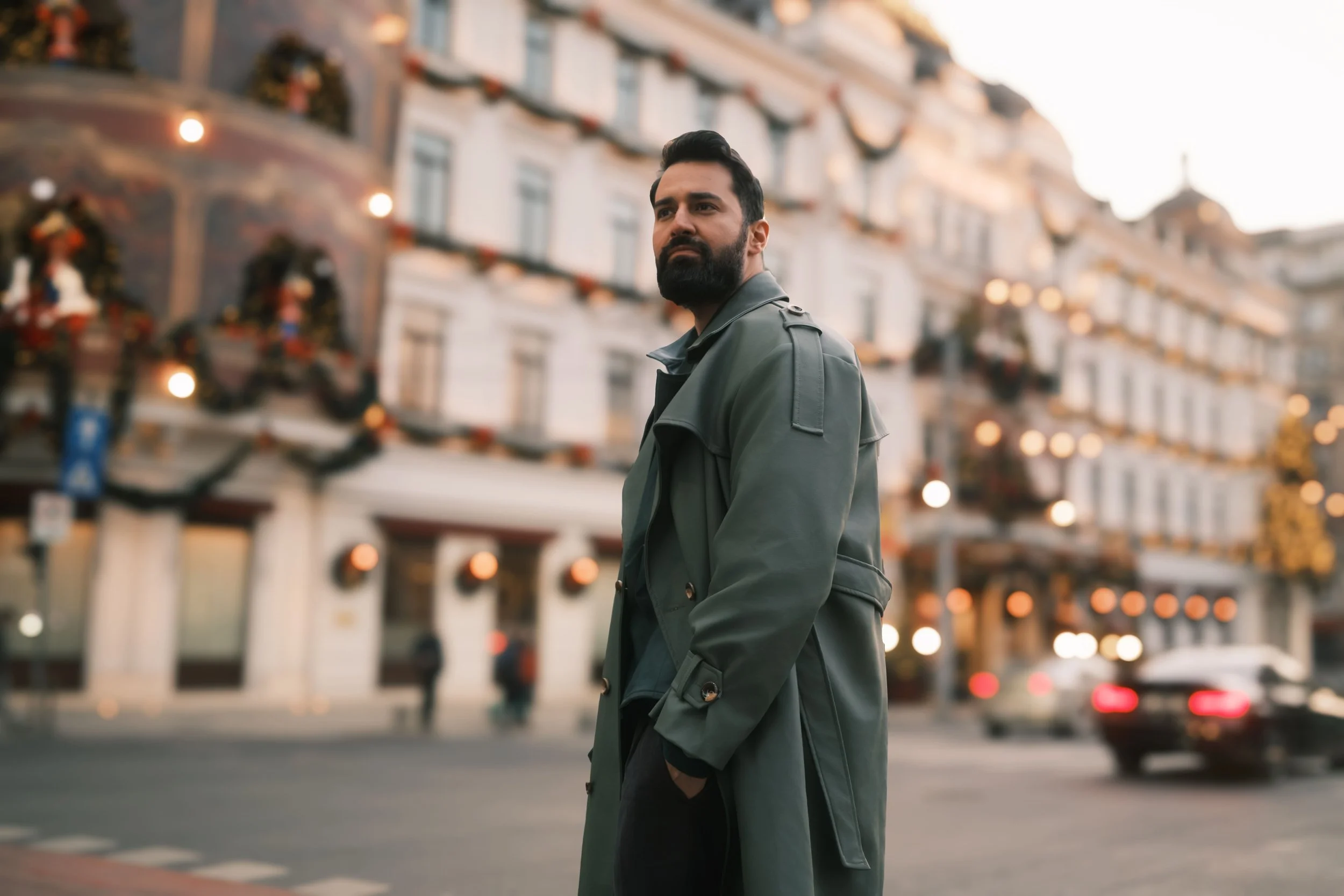 Ahmed Hatem on a city street at dusk in Bucharest, wearing a green trench coat, surrounded by soft lights and passing cars.