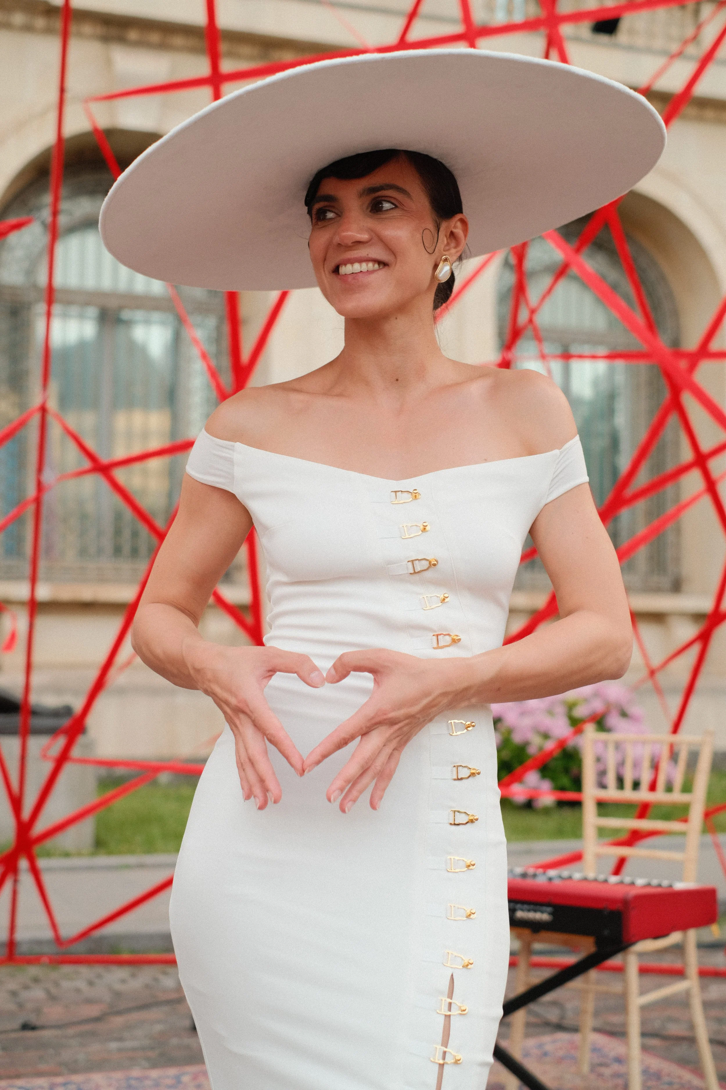 A woman wearing a white off-shoulder dress and a large wide-brimmed white hat, smiling and making a heart shape with her hands, in front of a decorative red geometric structure outdoors.