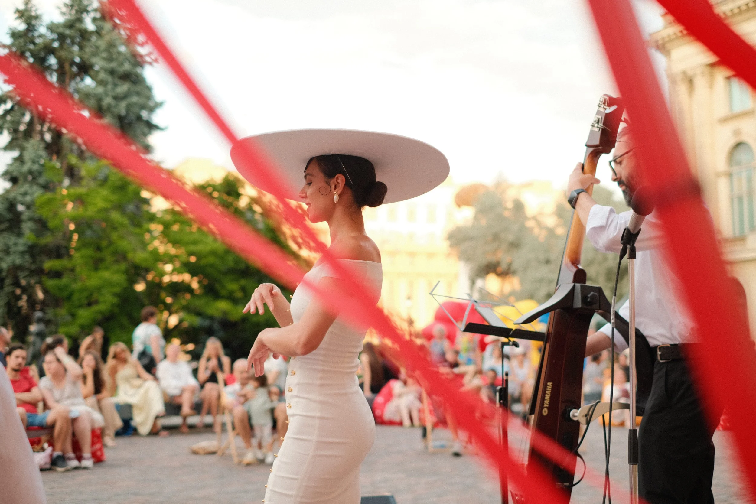 A woman in a white dress and large white hat dancing outdoors during a public event, with a musician playing a string instrument nearby, and a crowd seated and standing in the background.