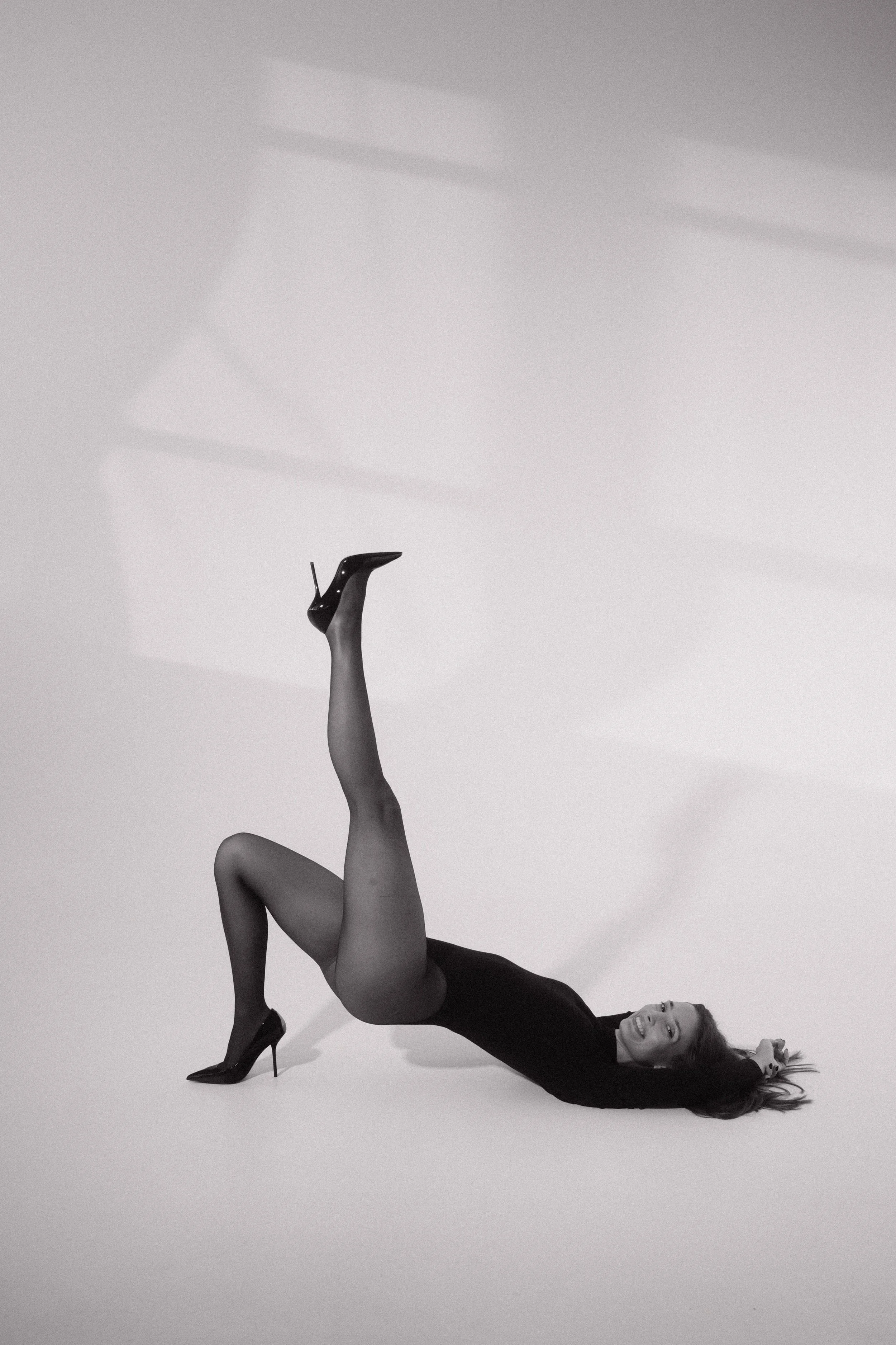 A woman in high heels and tights lying on the floor with one leg raised upward, smiling at the camera.