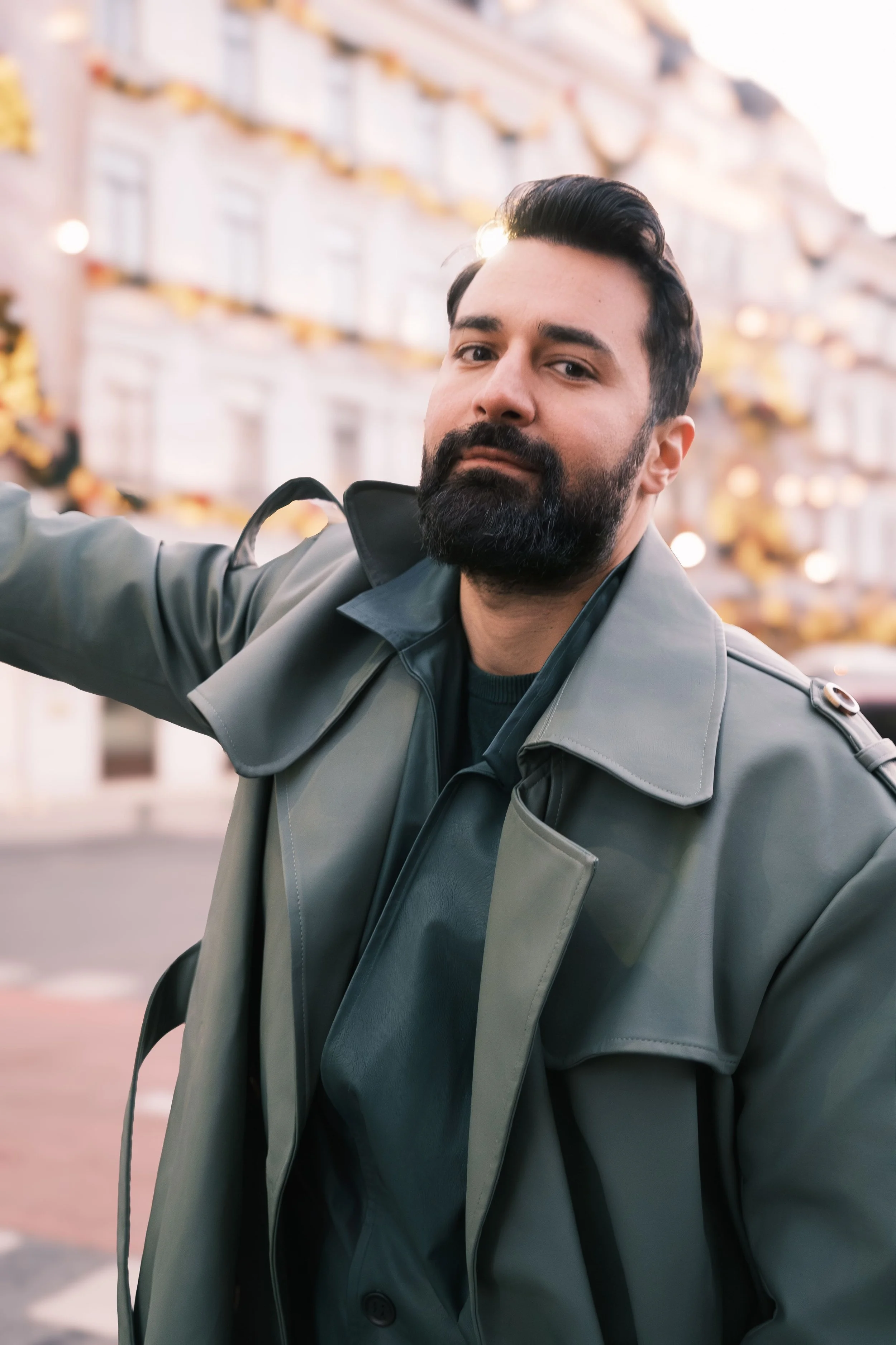 Ahmed Hatem taking a selfie in a quiet urban setting, wearing a gray trench coat, with soft lights and blurred architecture in the background.
