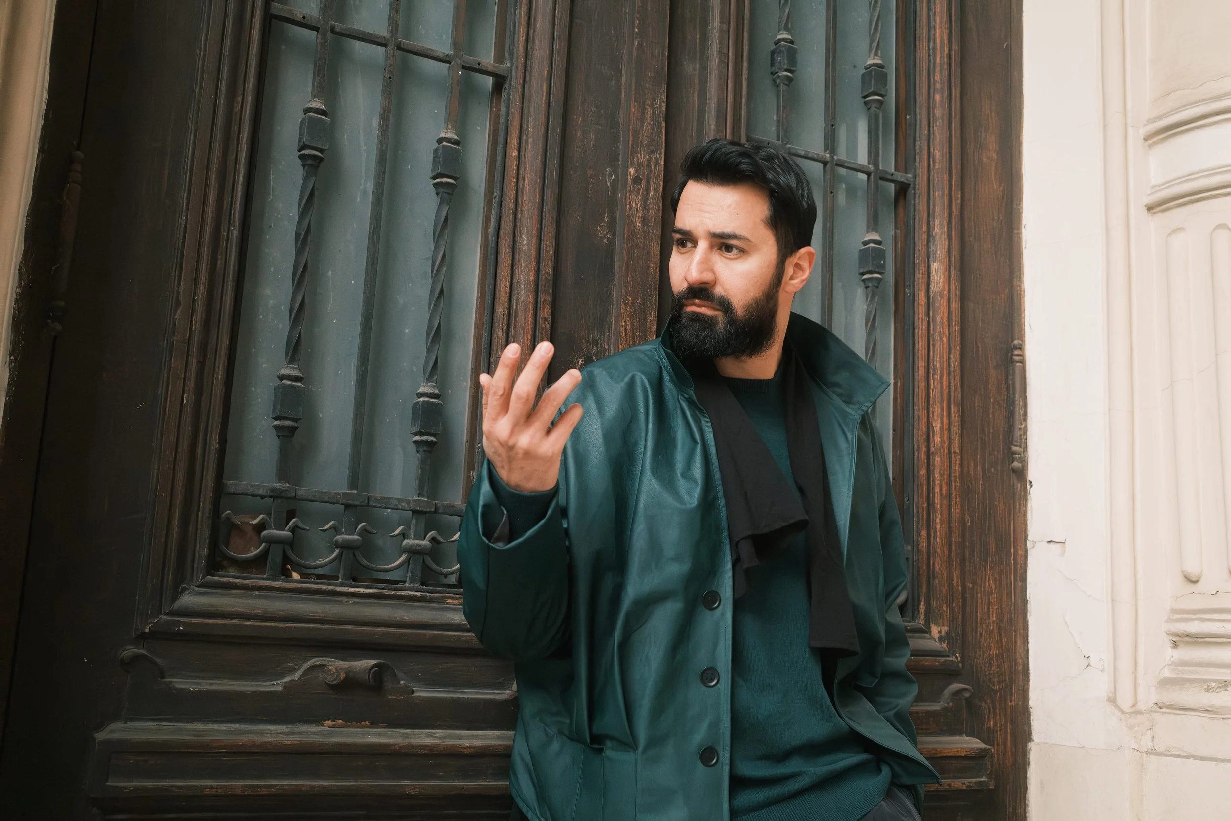 A portrait of Ahmed Hatem near a wooden window with iron bars, wearing a teal jacket, caught mid-gesture while looking away.