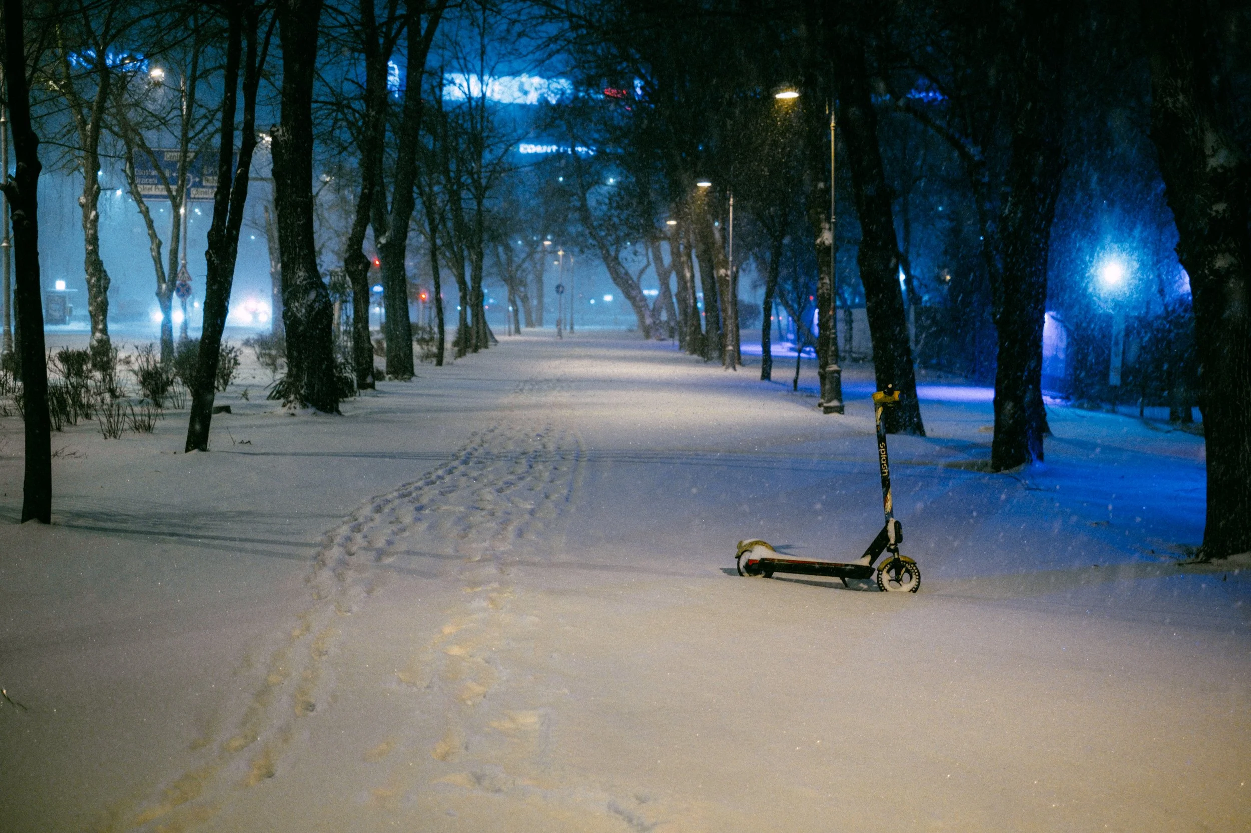 A snow-covered urban park at night with an electric scooter laying on its side in the snow and a trail of footprints leading into the distance. The park is lined with trees and illuminated by streetlights with a blue hue, with some city buildings vis