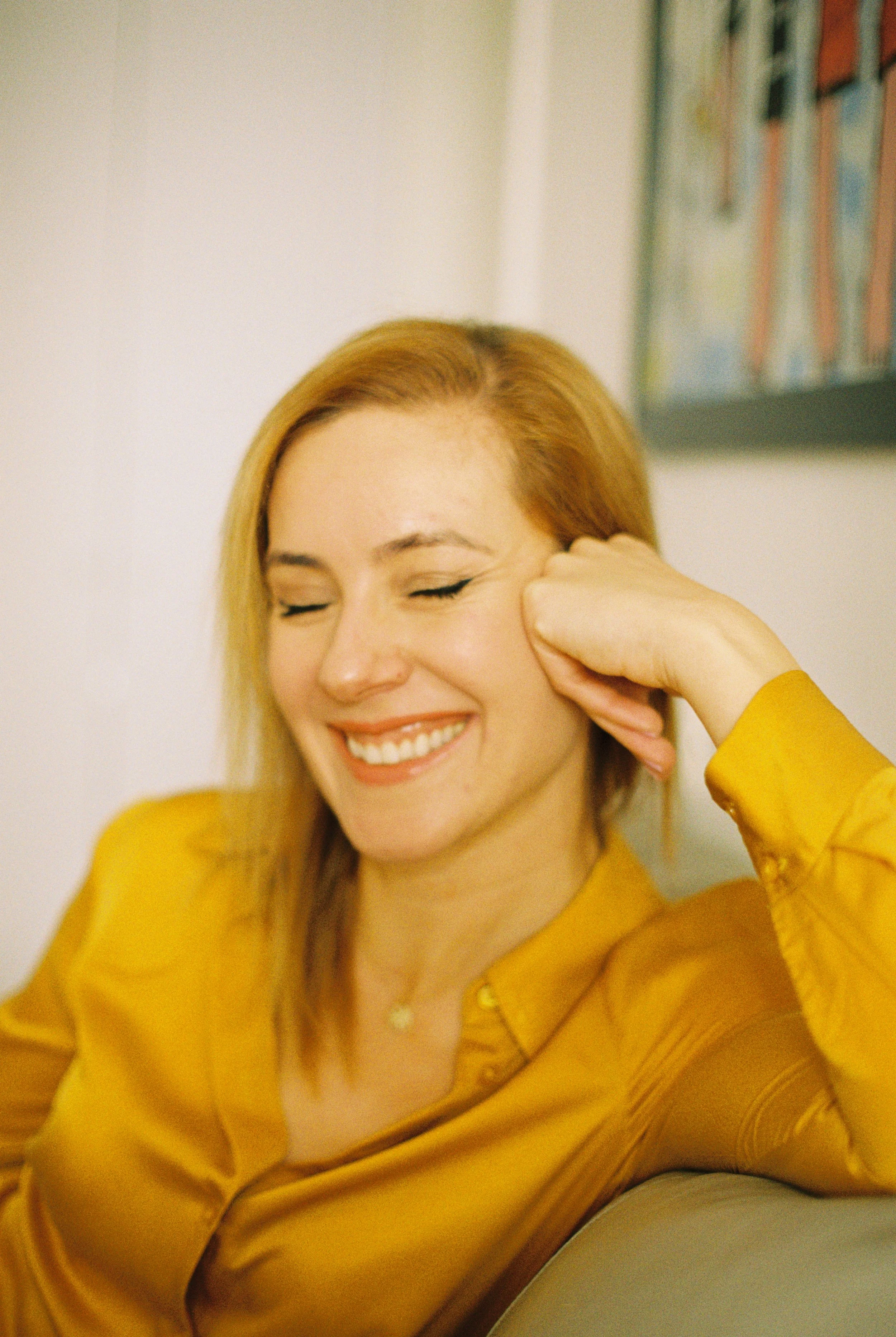 A woman with red hair, smiling and closing her eyes, wearing a yellow satin blouse, sitting indoors with a white wall and a colorful painting in the background.