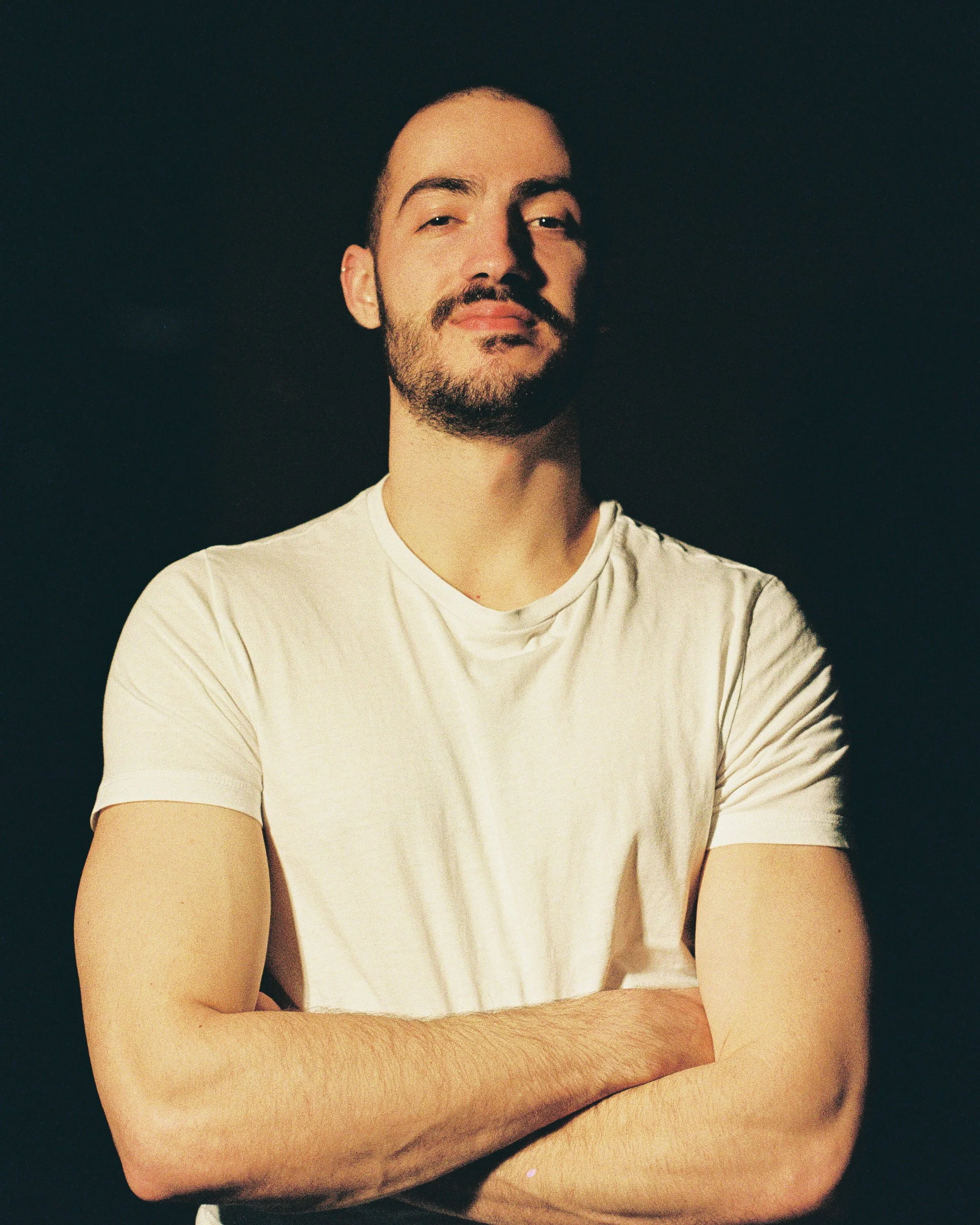 Portrait of a confident man with a beard and mustache, crossing his arms, wearing a white t-shirt, standing against a dark background.