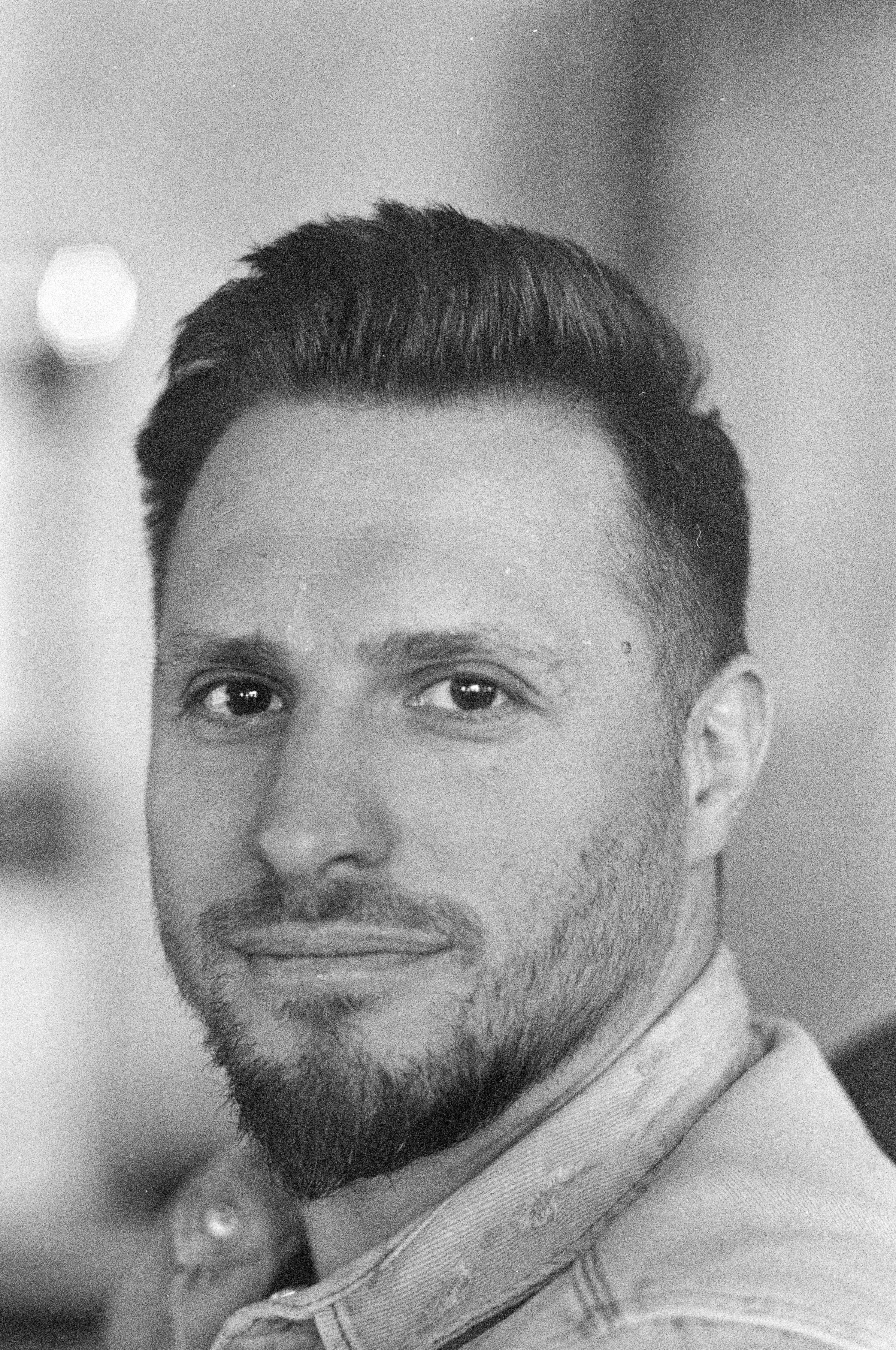 Black and white photograph of a young man with styled hair and a short beard, wearing a collared shirt, looking directly at the camera.