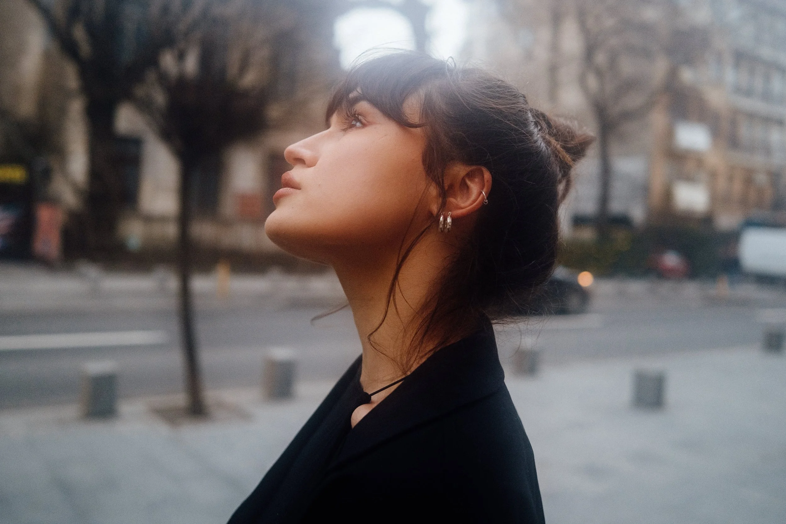 Side profile of a young woman with short dark hair, earrings, and a black jacket, standing outdoors in an urban setting with blurred buildings and trees in the background.