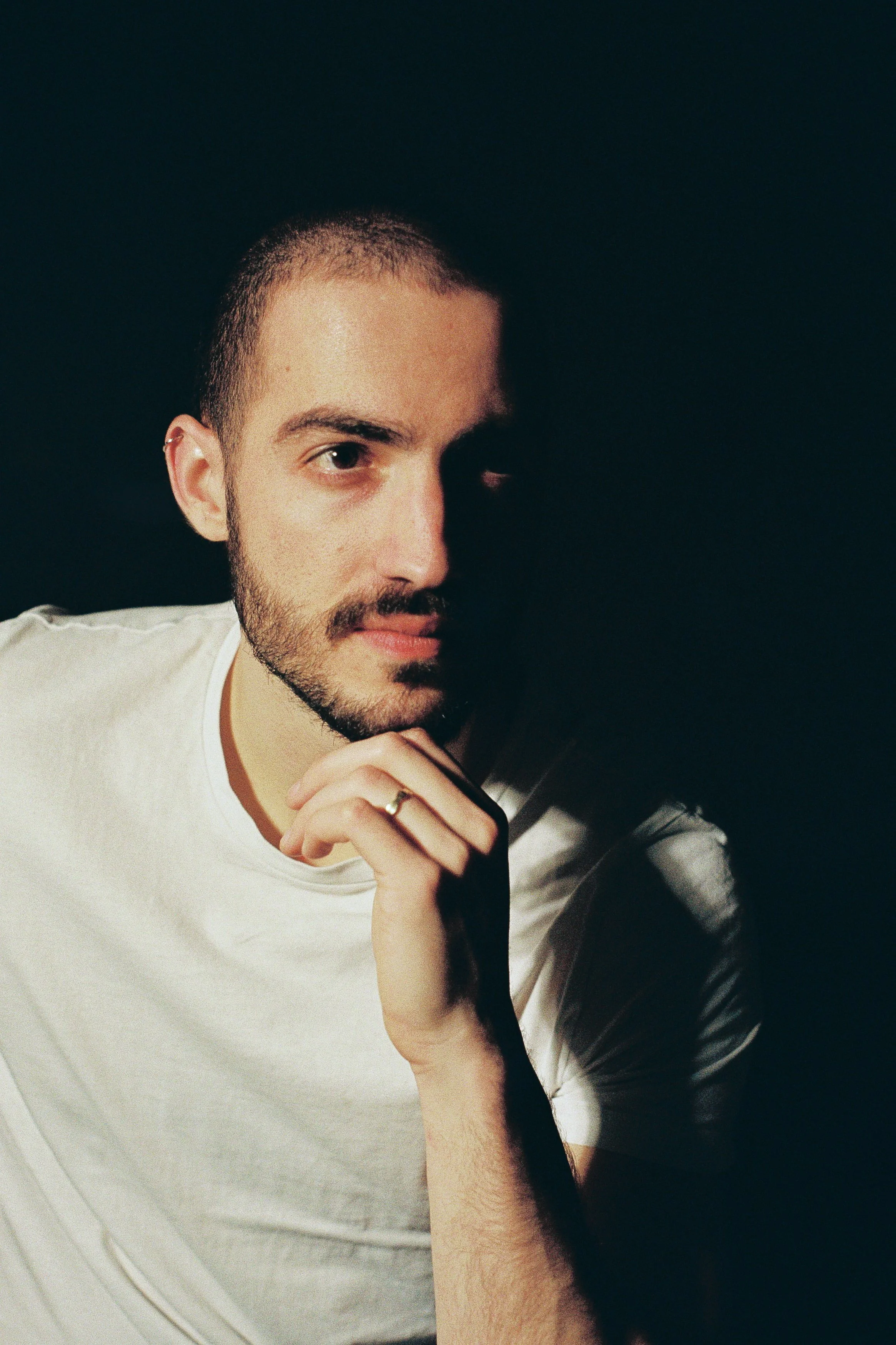 A young man with a beard and short hair, wearing a white t-shirt, is holding his chin with his hand and looking directly at the camera against a dark background.