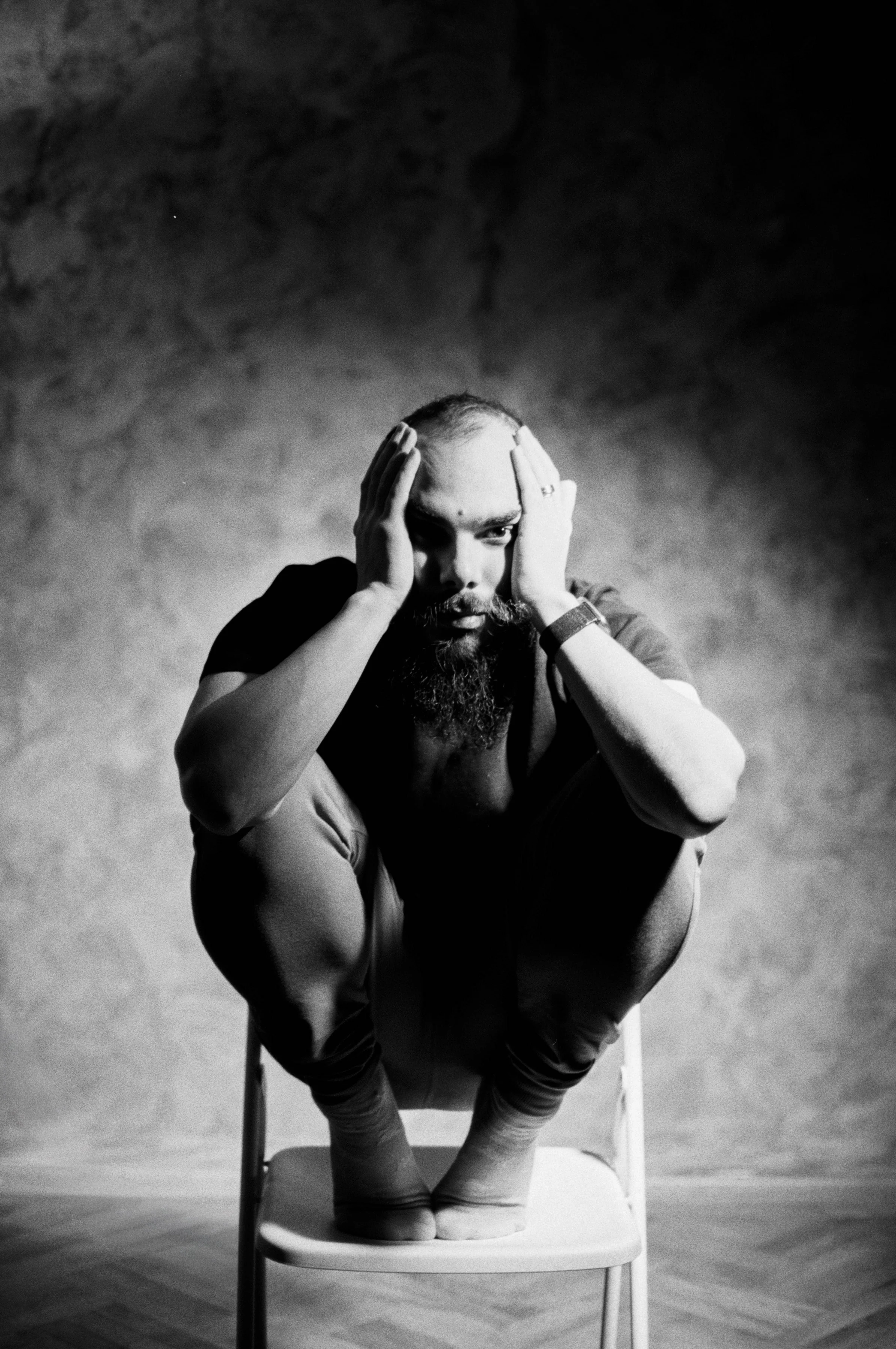 A man with a beard and mustache sitting on a chair with his knees drawn up, holding his head with both hands, in a contemplative or distressed pose in black and white.