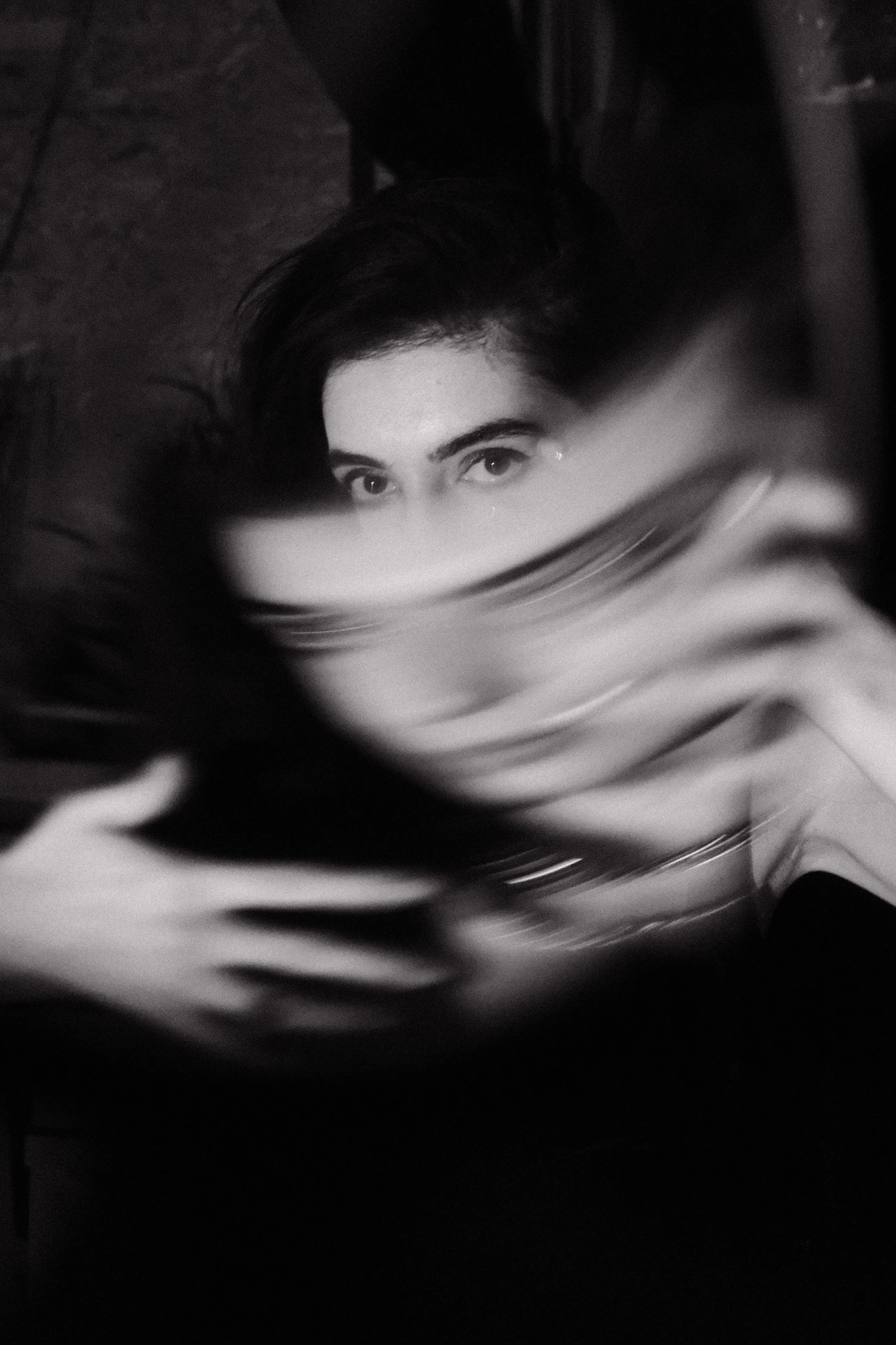 A black and white close-up photo of a woman lying on her back, with her face partially covered by a striped blanket or clothing. Only her eyes and part of her forehead are clearly visible, looking directly at the camera.
