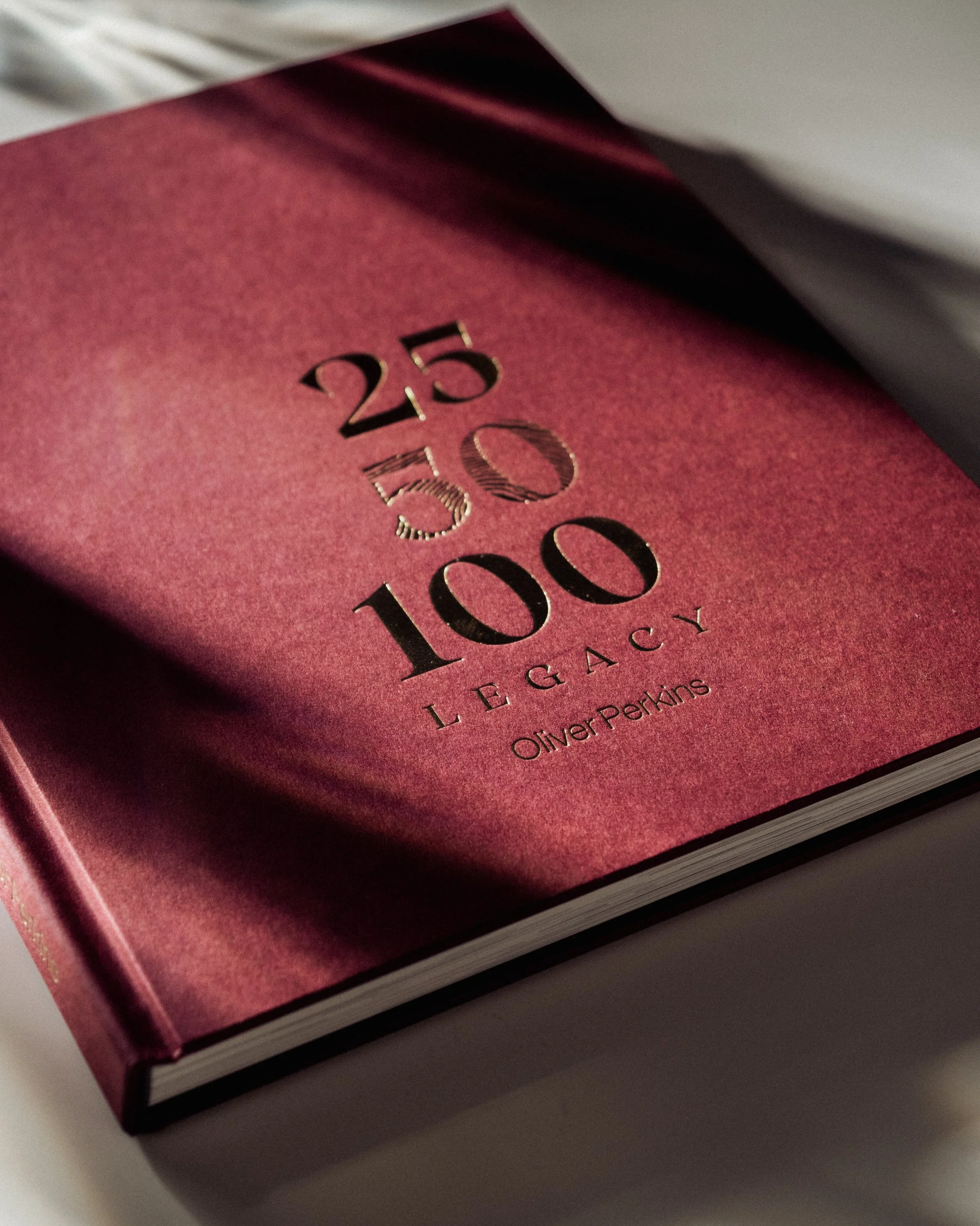 Close-up of a hardcover book titled '250 50 100 Legacy' by Oliver Perkins, with a red and black cover design.