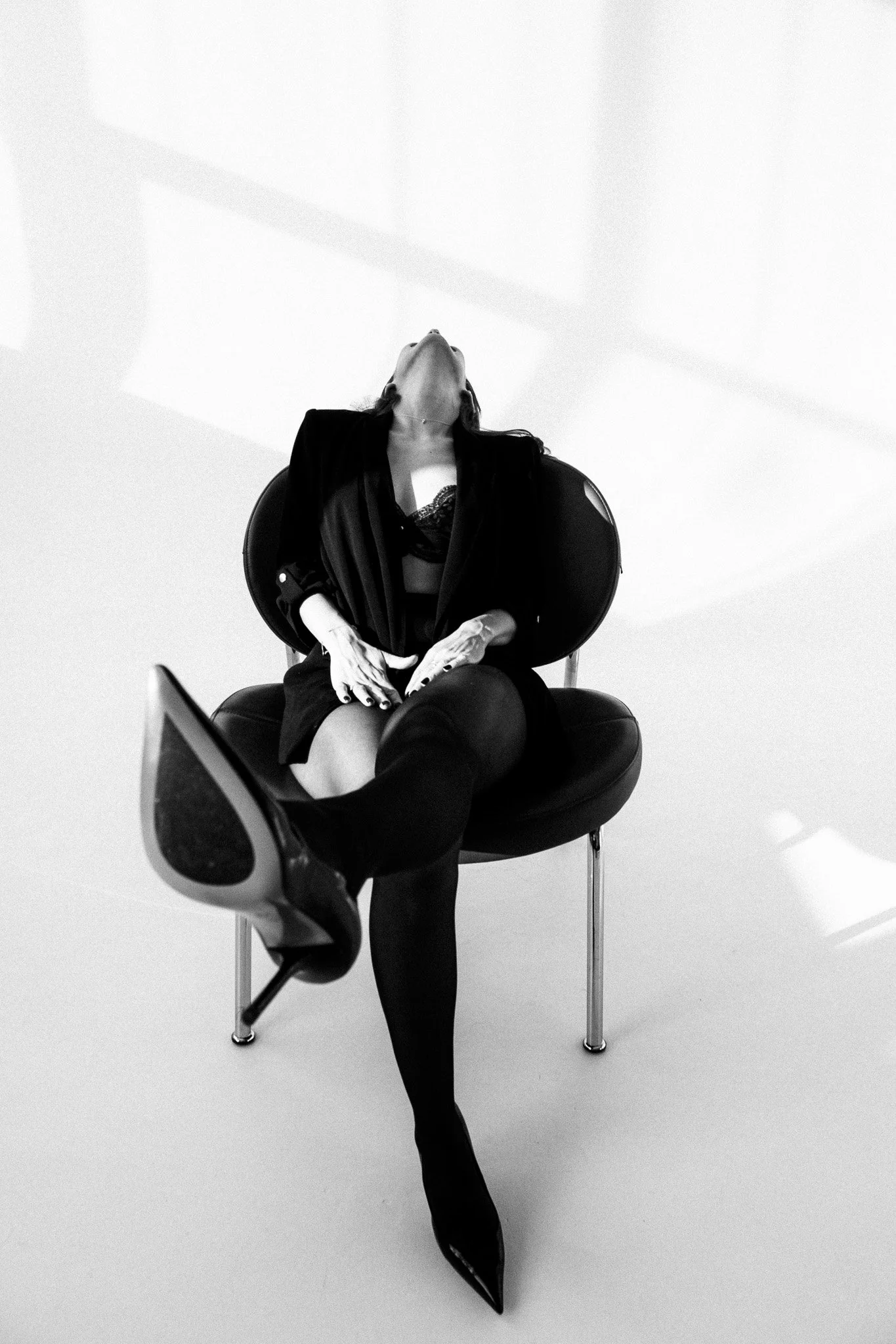 Black and white photo of a woman seated on a modern chair, her head tilted back, wearing a dark outfit with thigh-high stockings and high heels, with minimalist background and shadows on the wall.