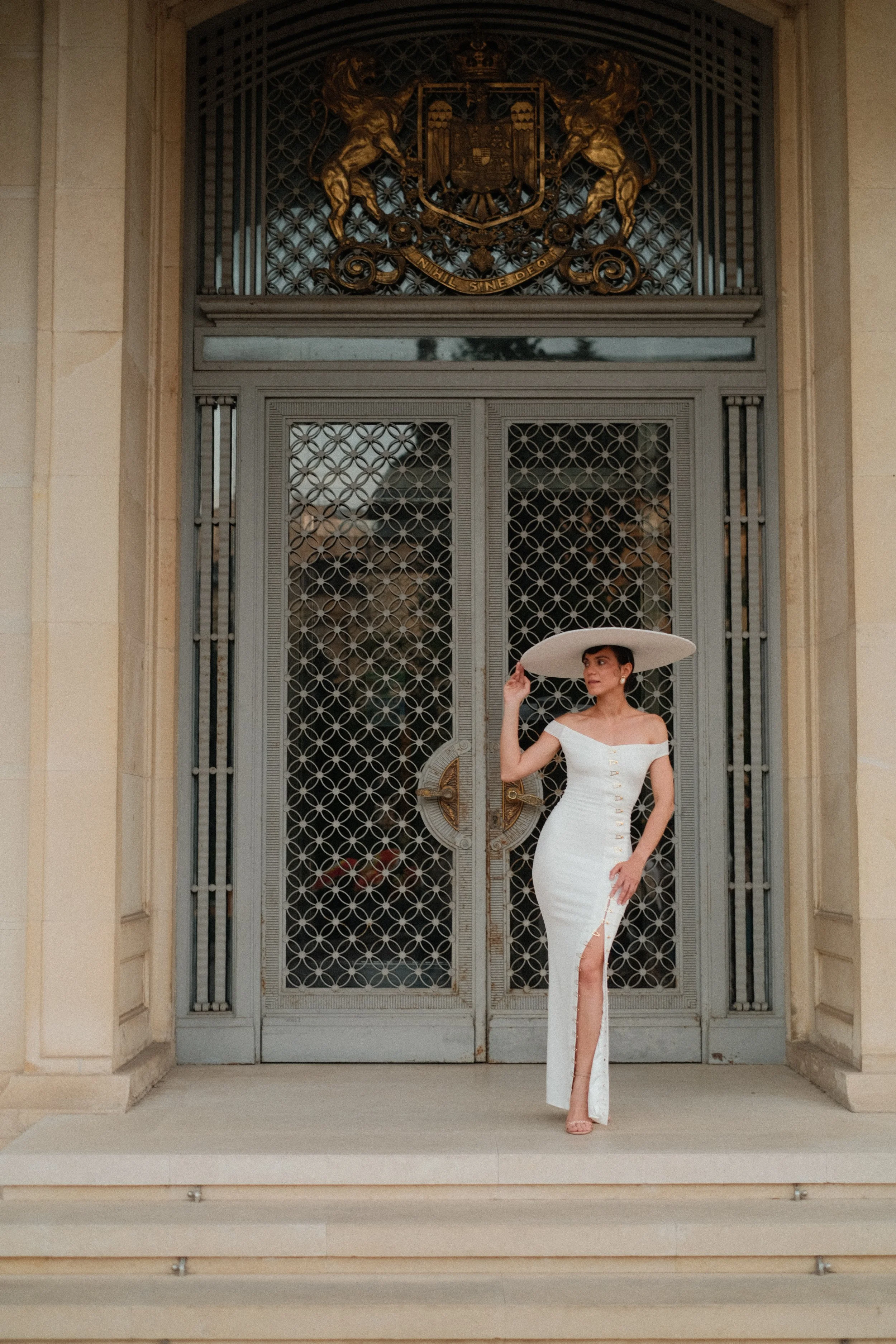 A woman in a fitted white dress with a slit and gold buttons, wearing a large white hat, stands in front of ornate metal doors with a crest above them, on the steps of a grand building.
