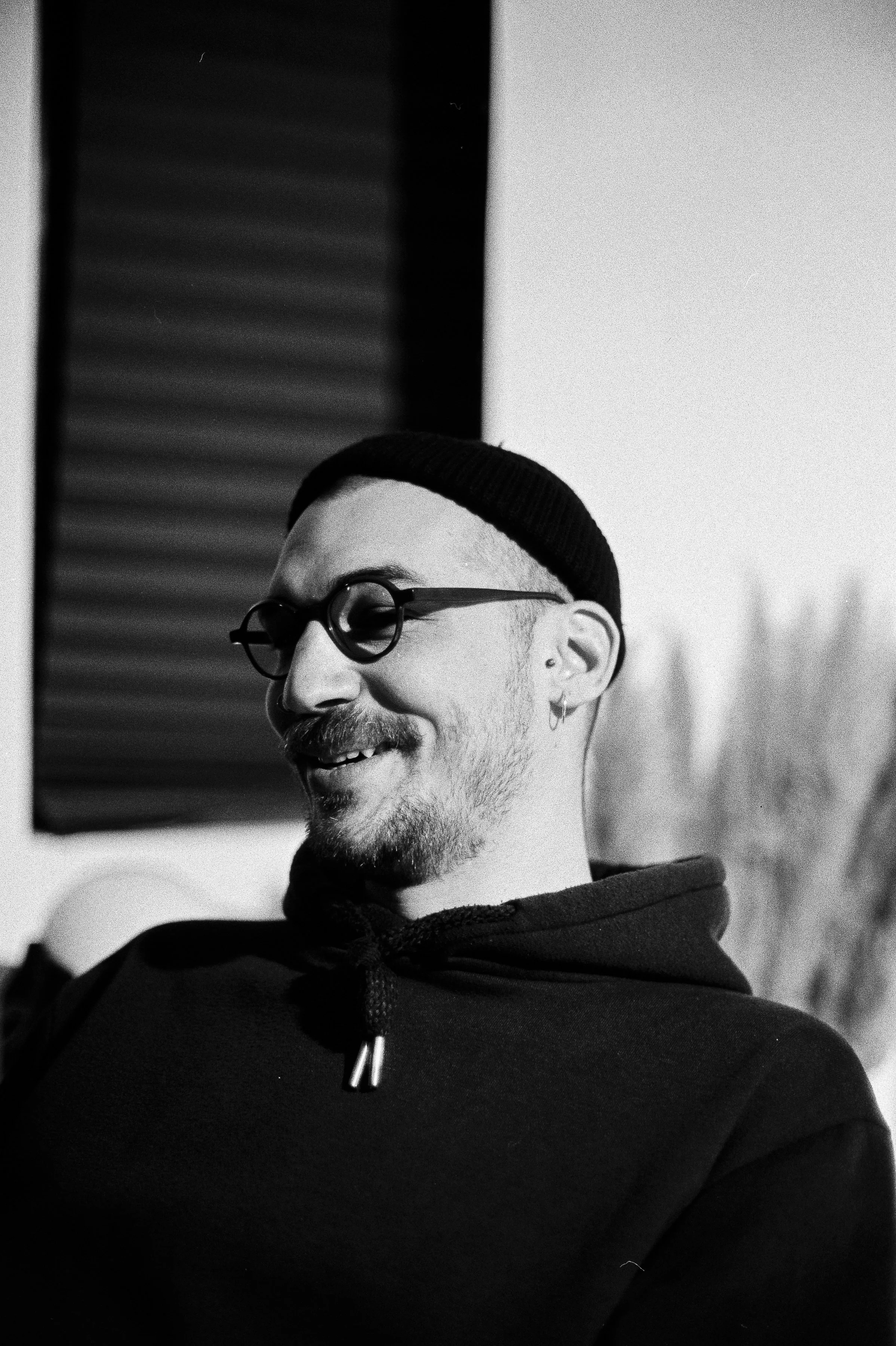 A black and white photo of a smiling man with glasses, a beanie, earrings, and a beard, sitting indoors.