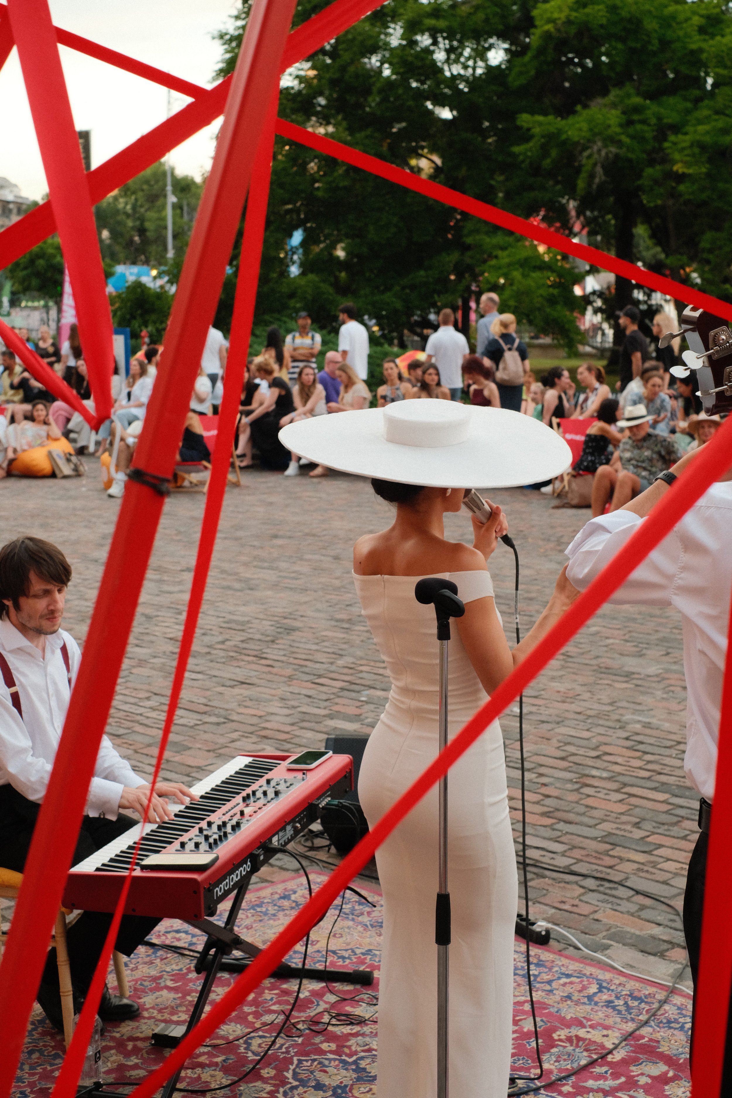 Musicians performing outdoors at a park, with a woman wearing a wide-brimmed white hat playing a microphone, a man playing a red keyboard, and people sitting and standing around, surrounded by trees and red metal structures.