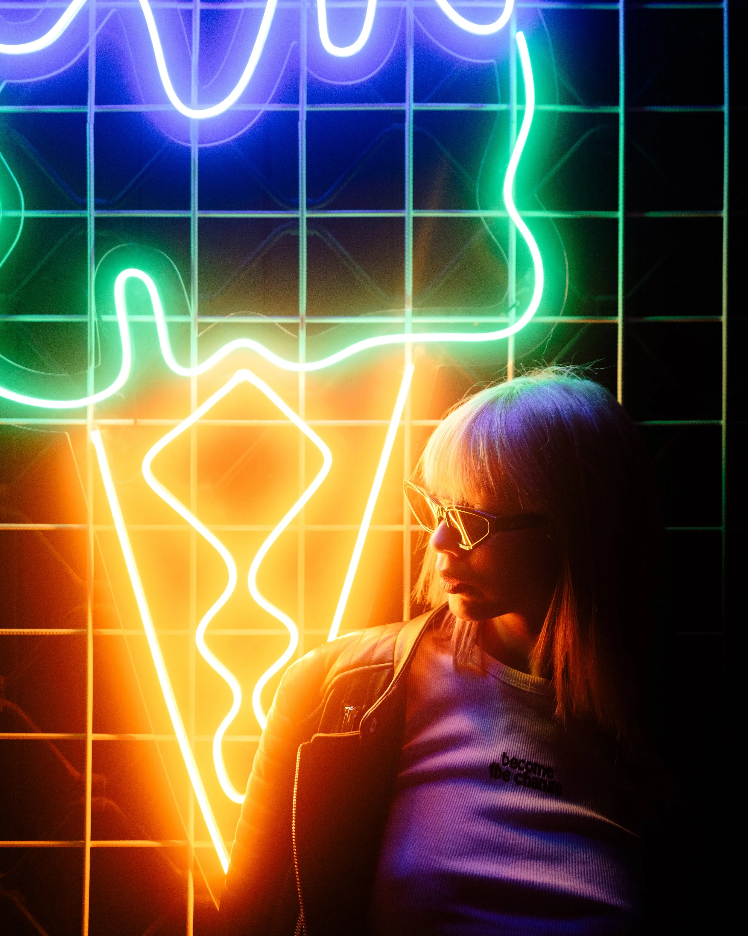 Person with blonde hair and sunglasses standing in front of a neon light display of an ice cream cone, with colorful glowing lights.
