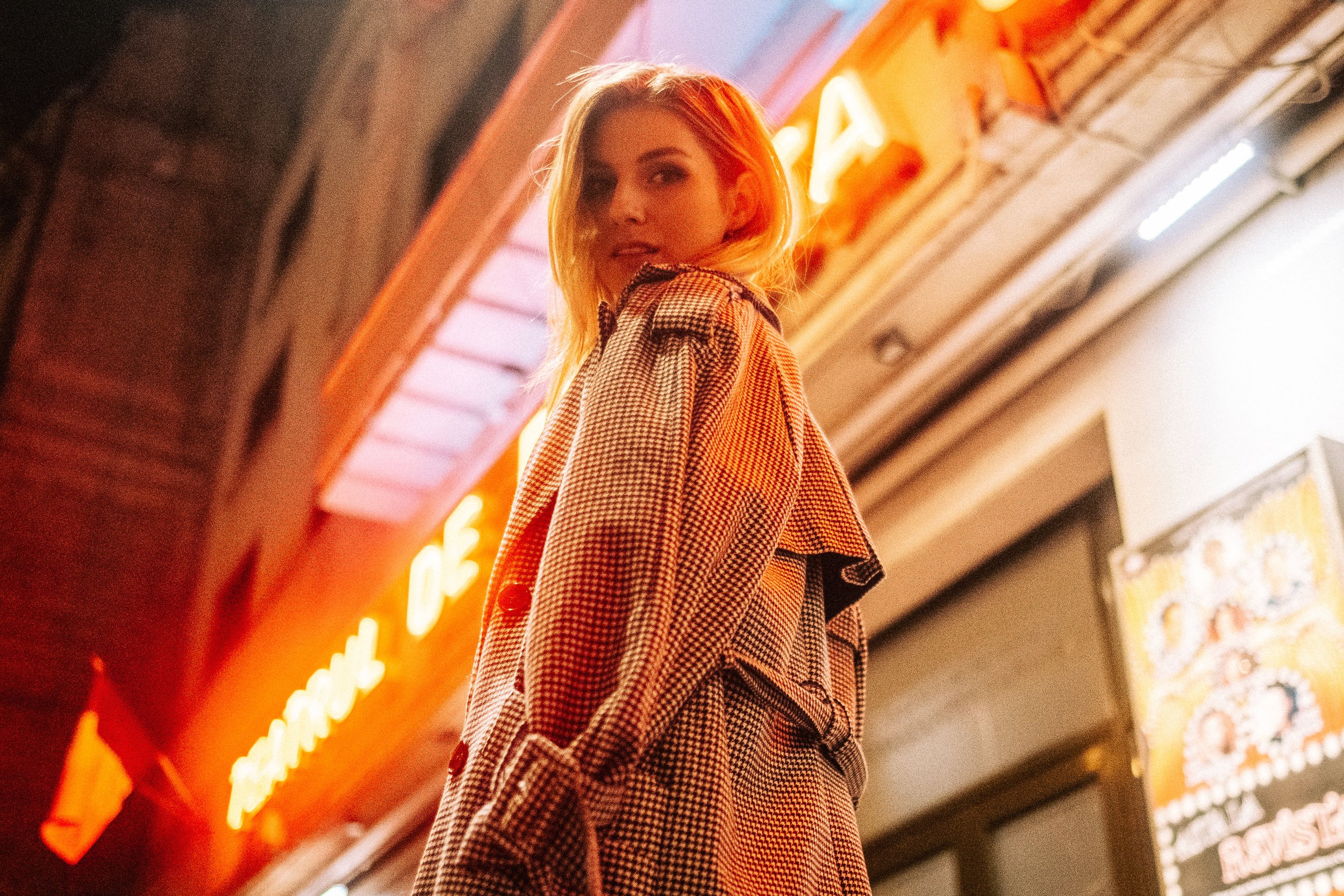 A woman with blonde hair wearing a checkered coat standing in front of a neon sign at night.