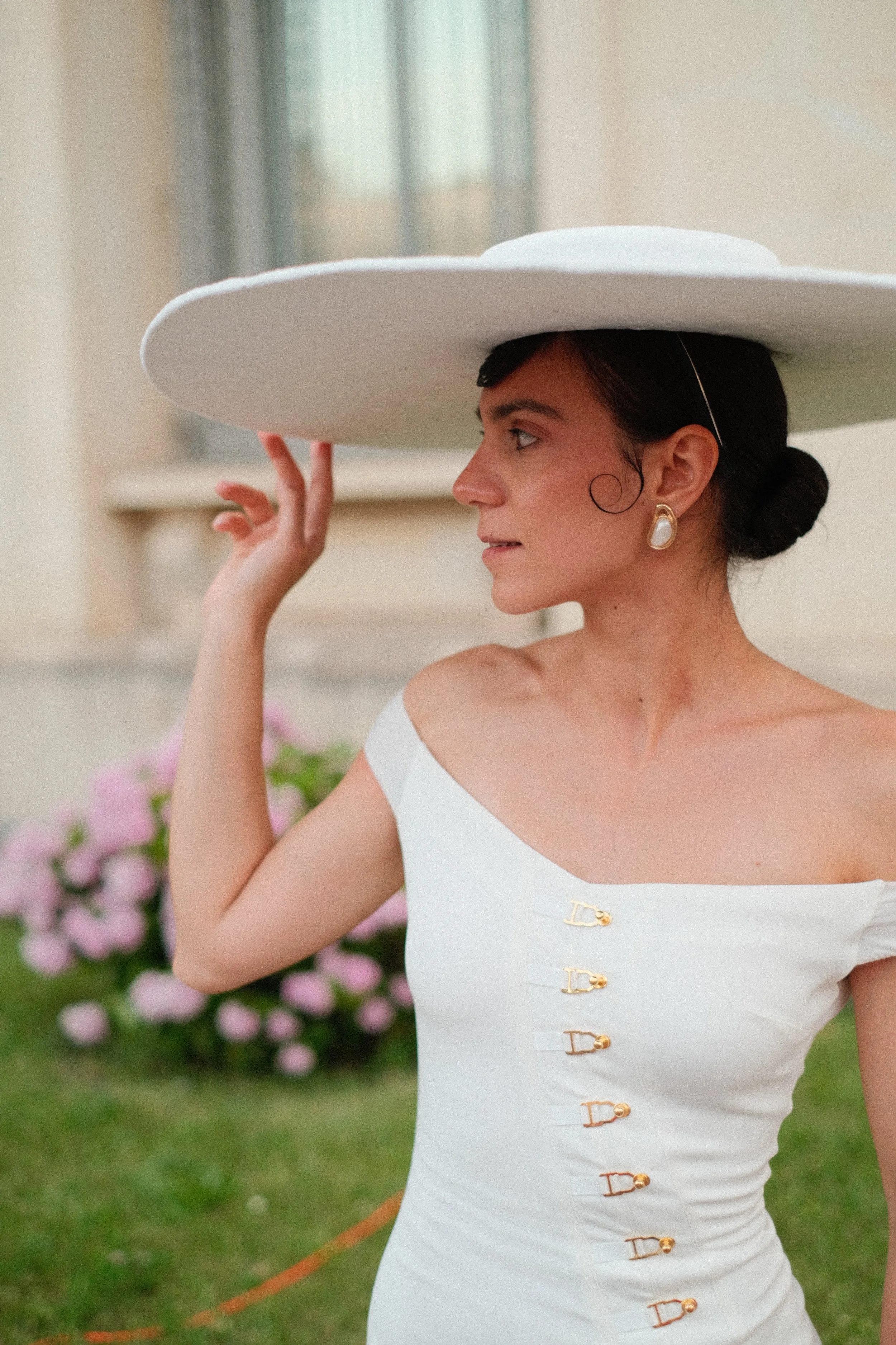 A woman in a white dress with gold buckles, wearing pearl earrings and a wide-brimmed white hat, standing outdoors in front of pink flowers and greenery.