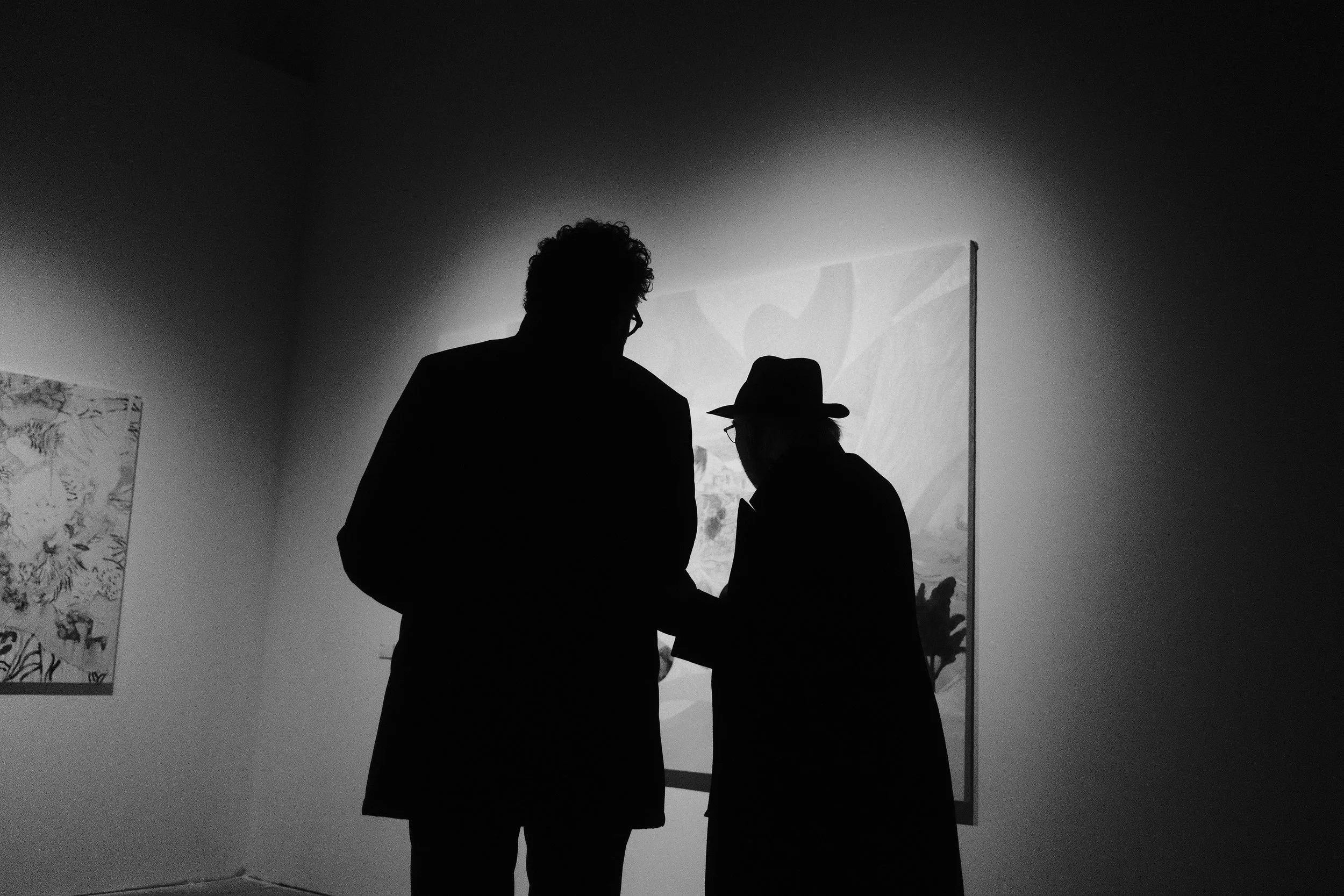 Silhouettes of two men having a conversation in an art gallery, with paintings on the wall behind them.