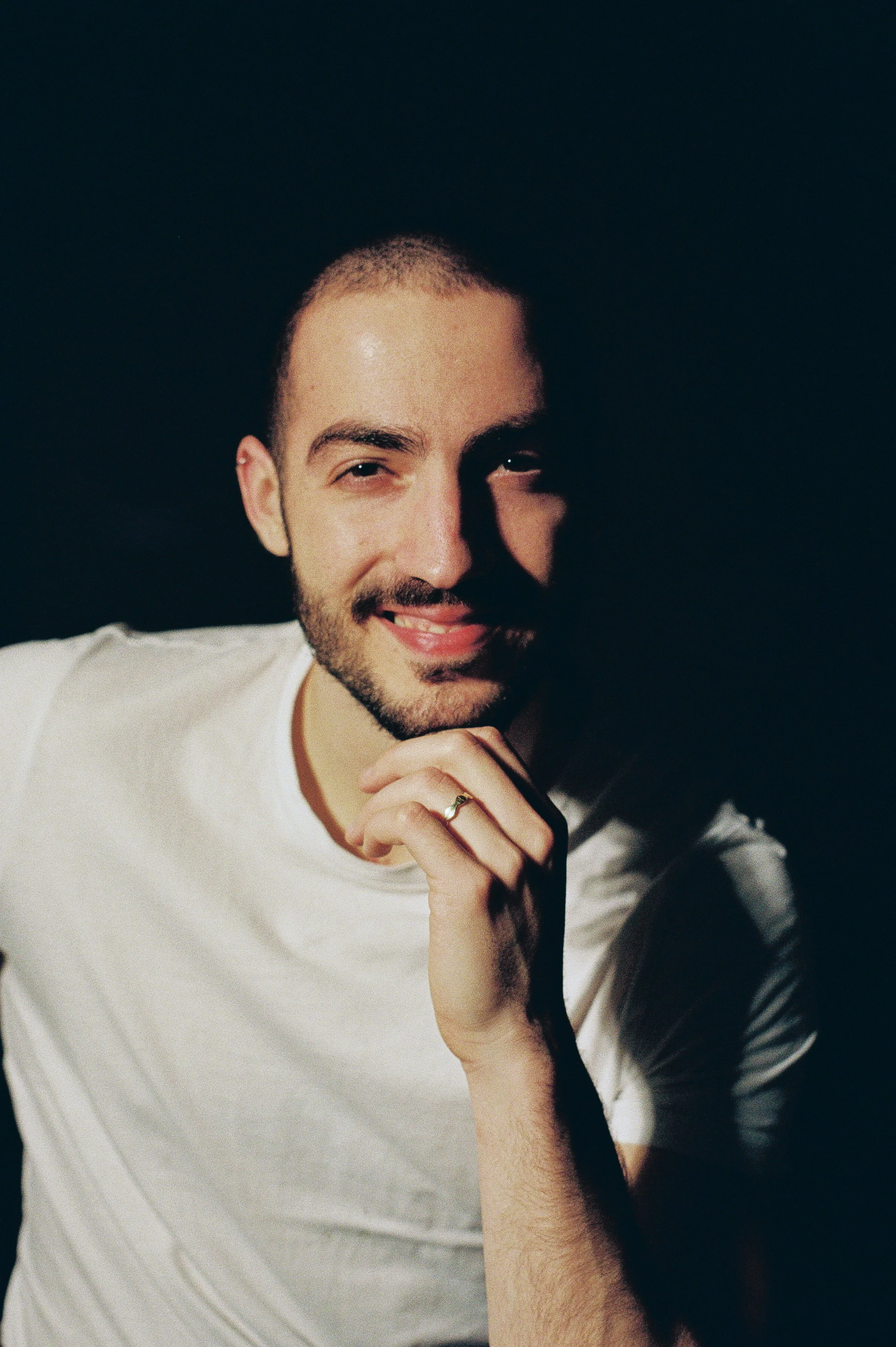 A young man with a beard and short hair smiling, with his hand resting on his chin, wearing a white t-shirt, in a dimly lit environment with a dark background.