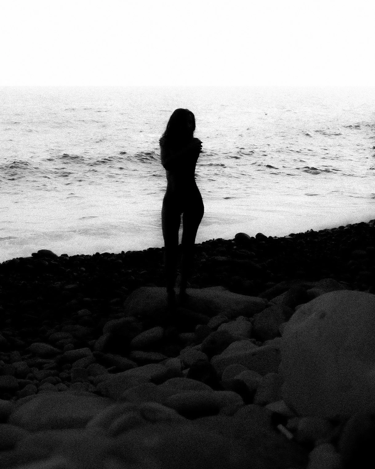 Silhouette of a woman standing on a rocky beach facing the ocean during sunset or sunrise.