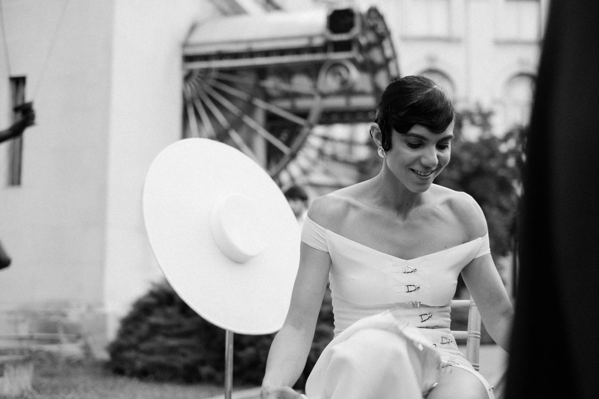 Black and white photo of a woman with short hair wearing an off-shoulder dress with button details, smiling, with large circular balloon in the background and a Ferris wheel in the distance.