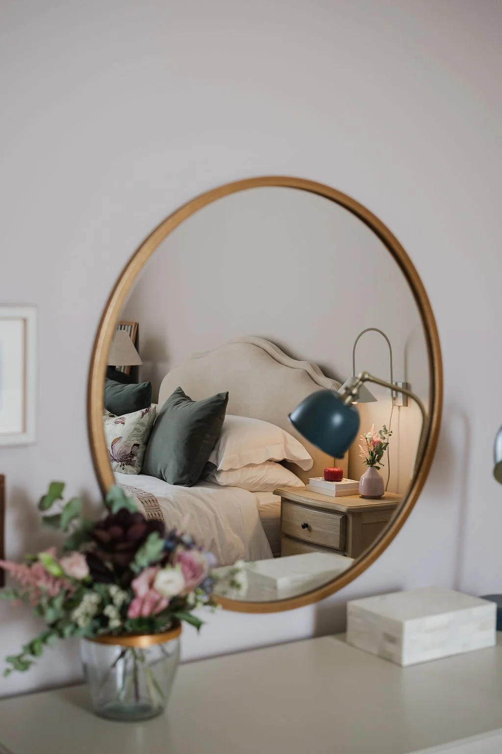 A bedroom reflected in a round mirror, featuring a bed with plush pillows, a bedside table with a vase of pink flowers, and a lamp on the wall. Kinross Edinburgh Scotland