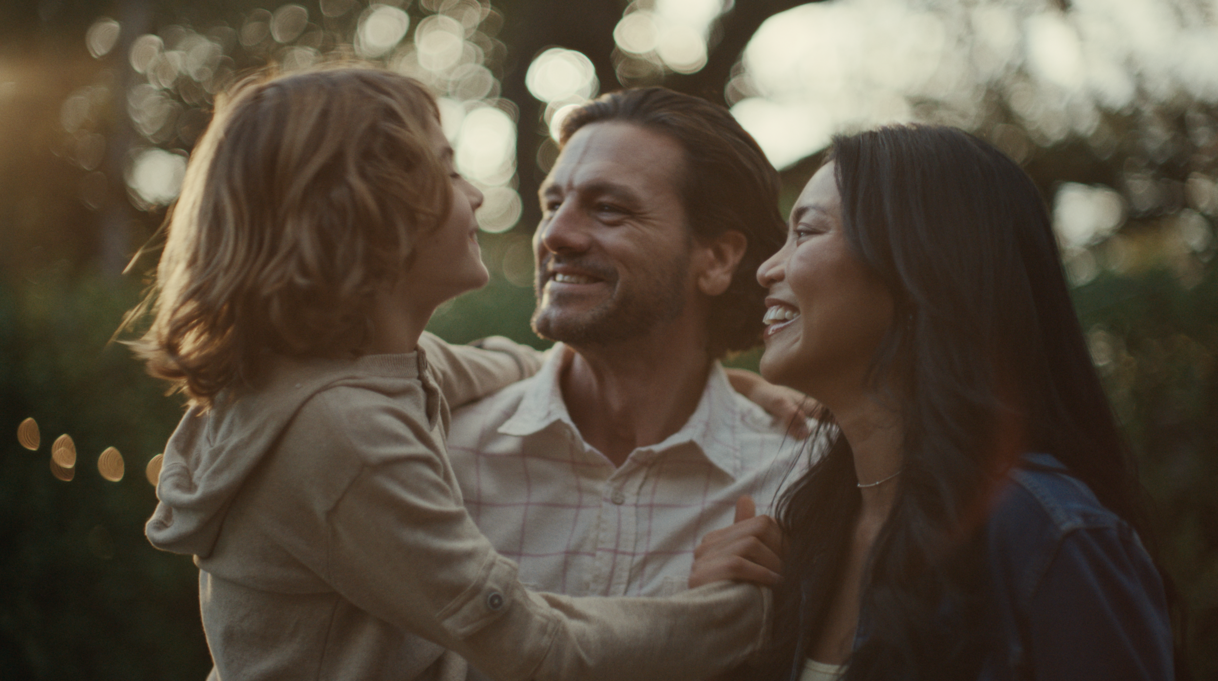 A family of three, consisting of a woman, a man, and a young child, enjoying each other's company outdoors during sunset. The woman and man are smiling and facing each other, while the child looks at the woman with affection. The background is softly blurred with bokeh effect from the sunlight filtering through trees.